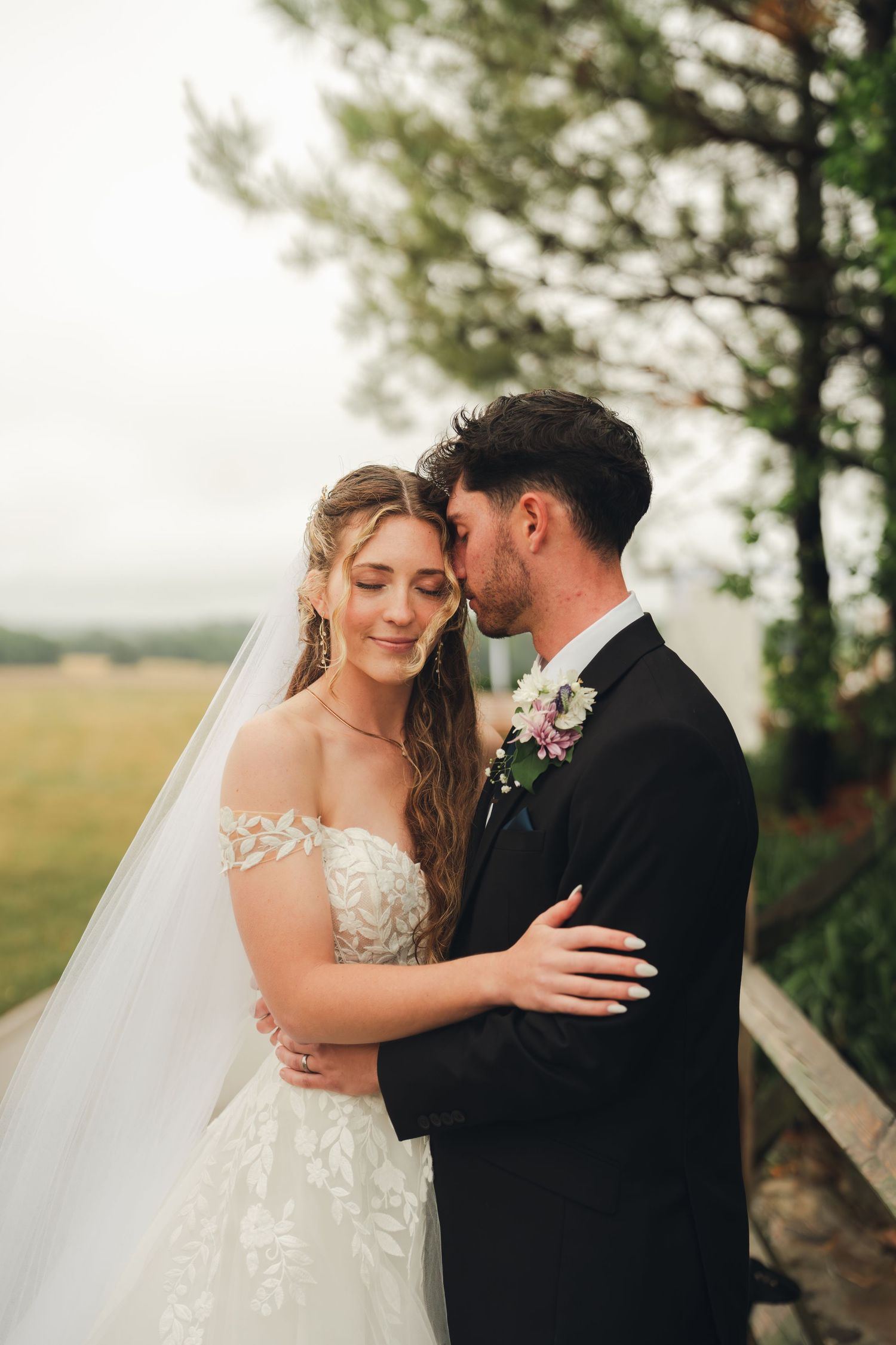 Groom kissing bride on forehead as she smiles in an off-shoulder lace gown in a field.