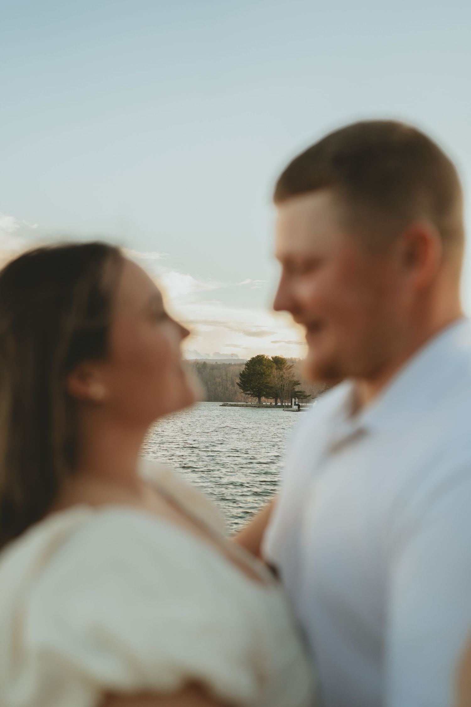 Romantic blurred couple facing each other with a serene lake and lone tree in the background.