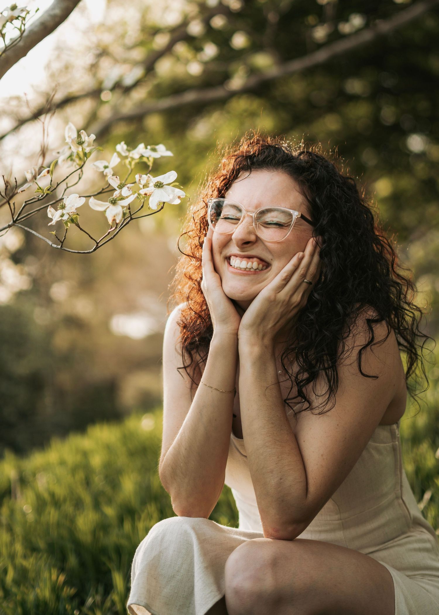 A laughing curly red-haired woman with glasses sits outdoors near white blossoms in warm golden light.