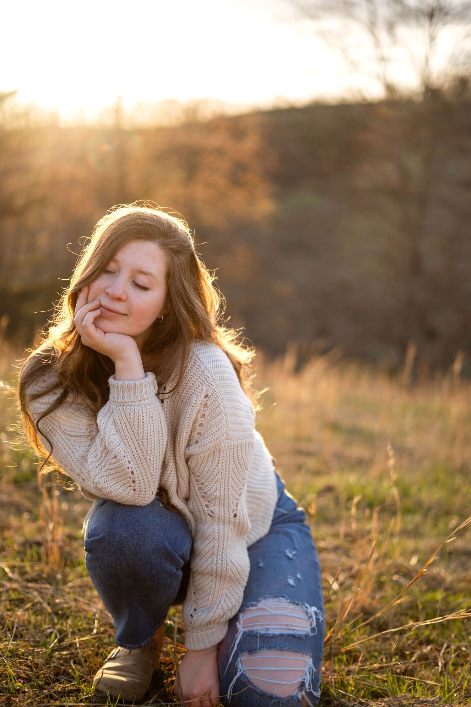 Teenage girl in beige knit sweater and ripped jeans sitting in sunlit meadow with eyes closed.