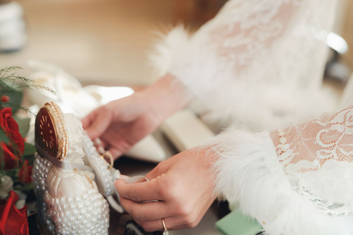 A bride in a lace robe with feather cuffs carefully ties her pearl-embellished white wedding shoes.