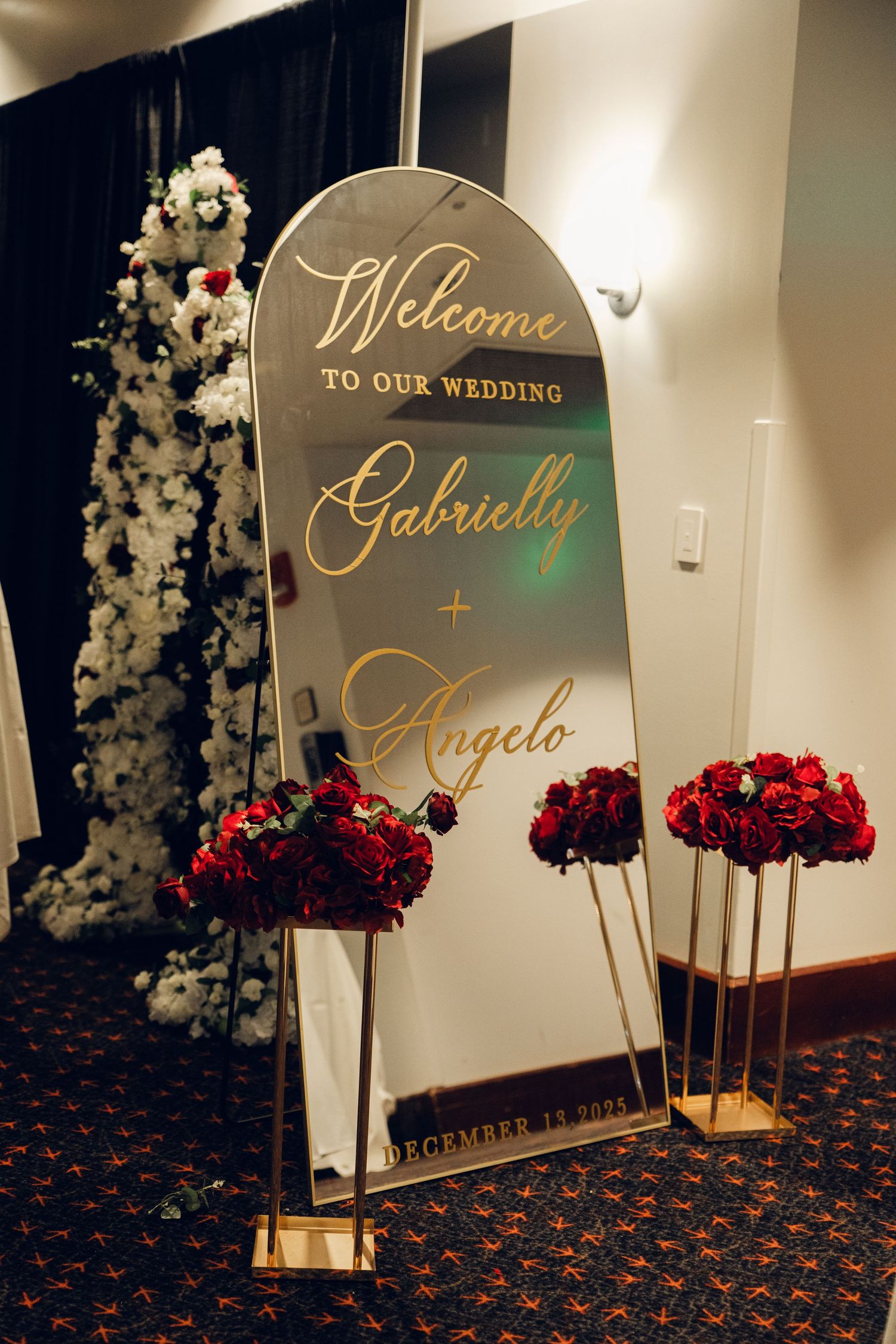 Gold mirrored wedding welcome sign for Gabrielly and Angelo surrounded by red roses and white floral arrangements.