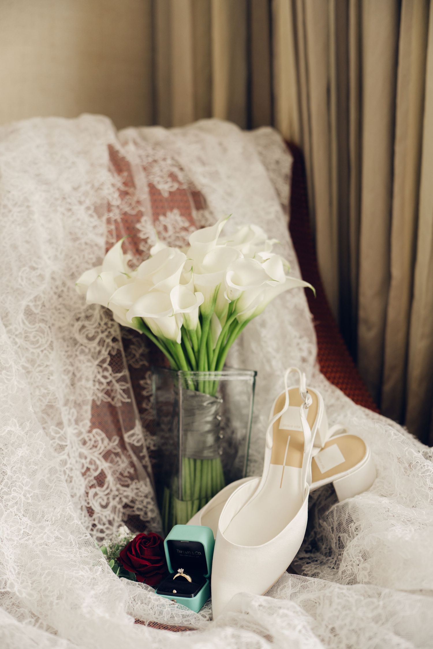White calla lily bouquet in vase with bridal heels, ring box, and lace veil on chair.