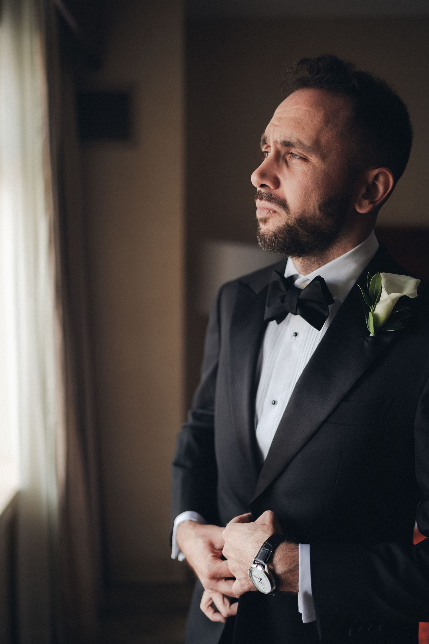 Elegant groom in black tuxedo with bow tie and boutonniere gazing out window on wedding day.