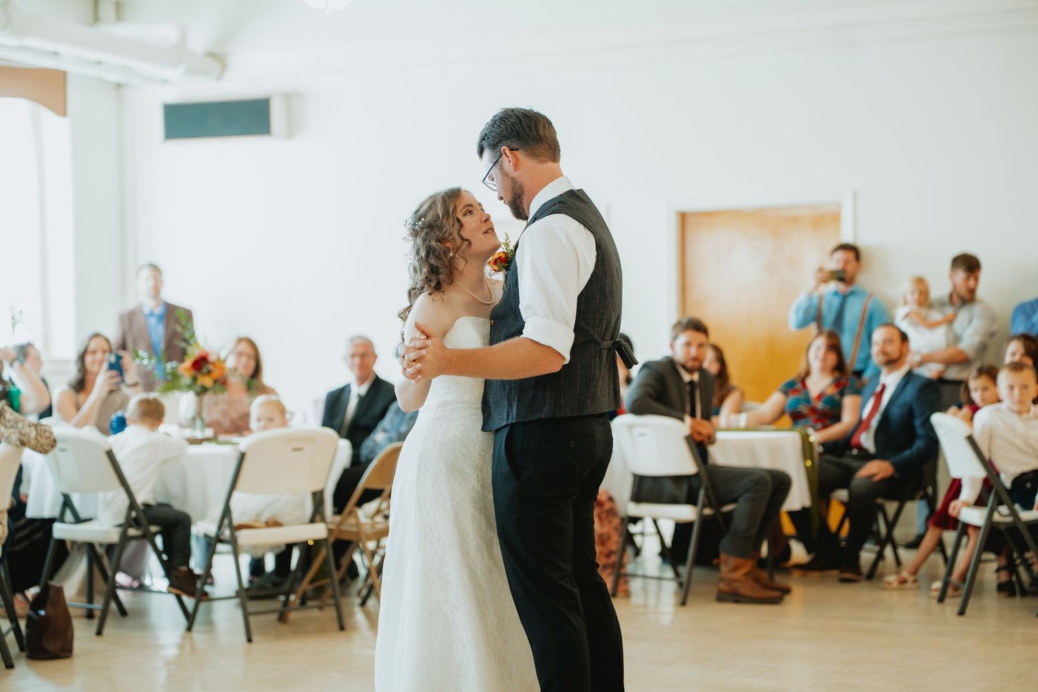 Bride and groom share their first dance at a wedding reception, surrounded by seated guests in a bright white venue.
