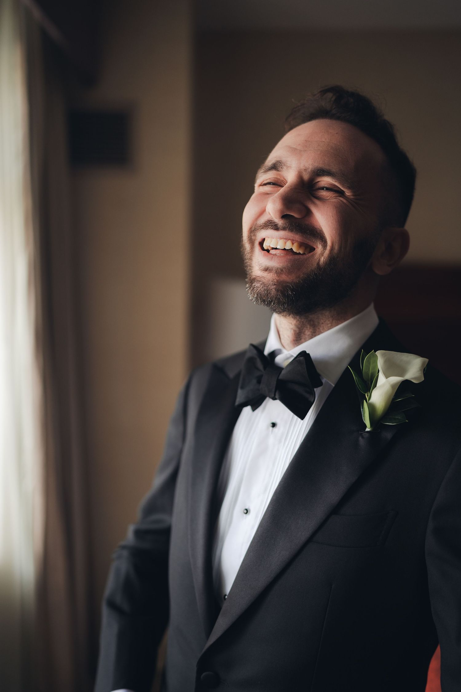 Joyful groom in black tuxedo laughing with eyes closed on wedding day by window.