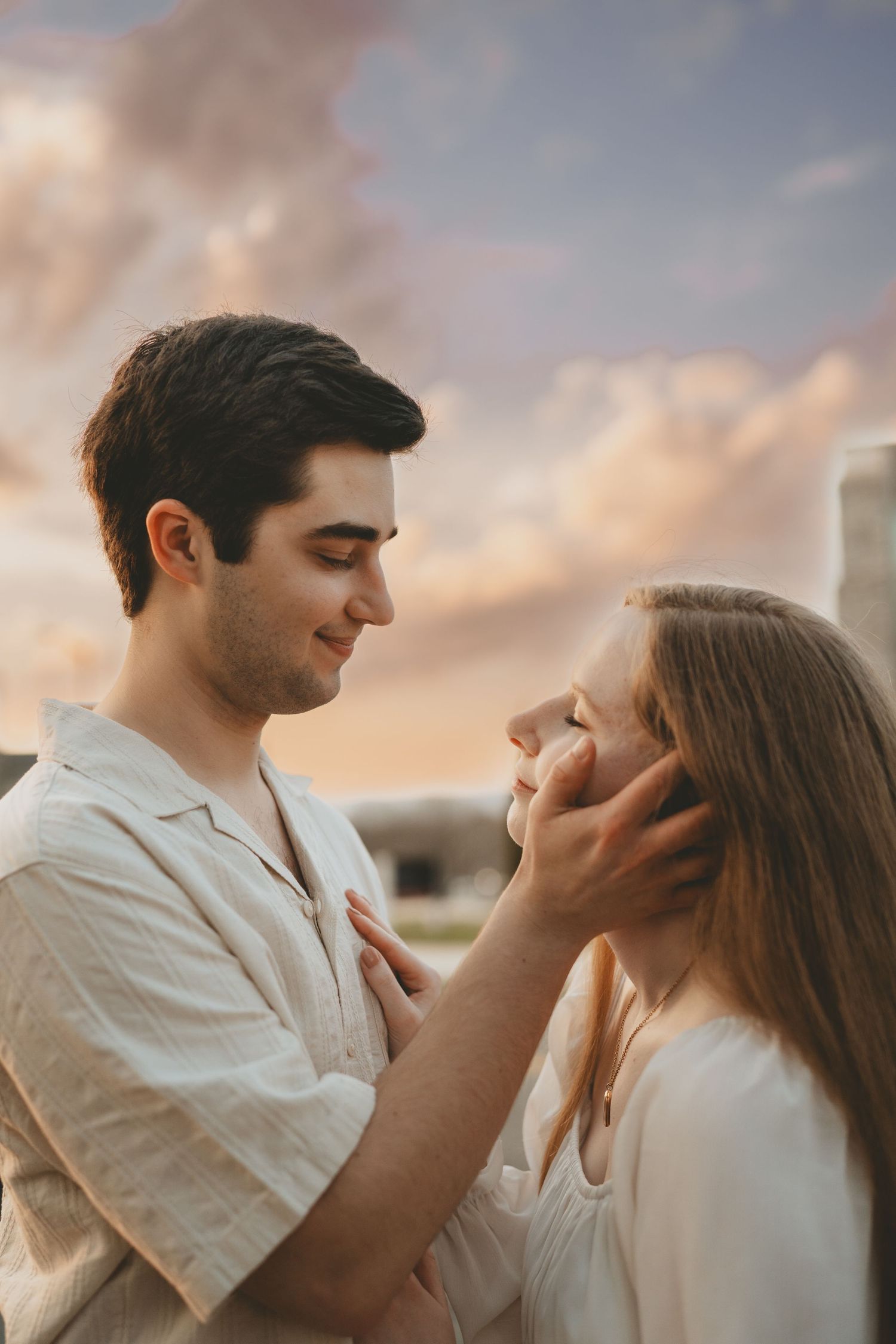 Young man tenderly cups womans face as they share an intimate moment under a pastel sunset sky.