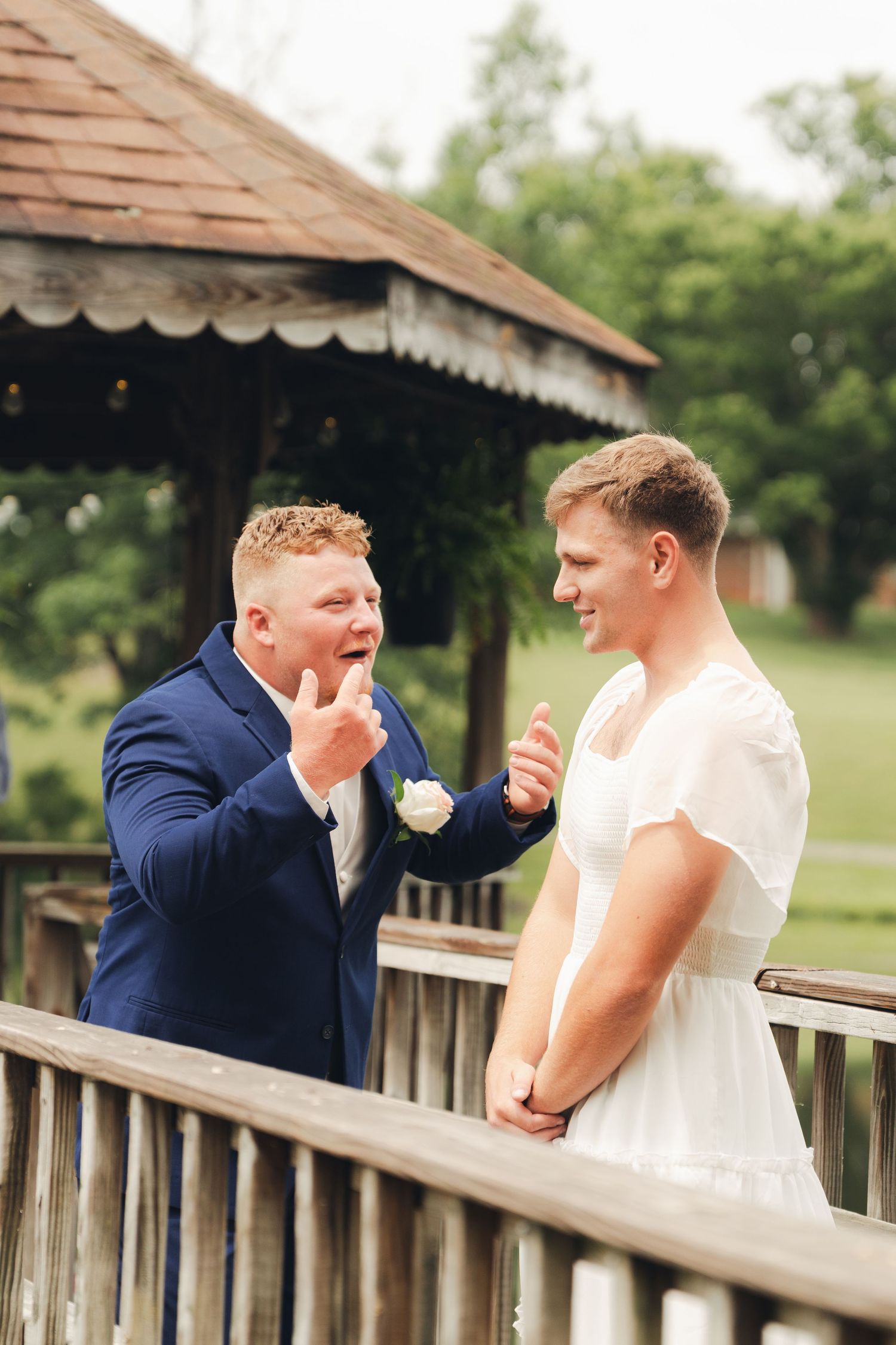 Groom in white dress and best man in blue suit share a joyful moment on a wooden deck at an outdoor wedding.