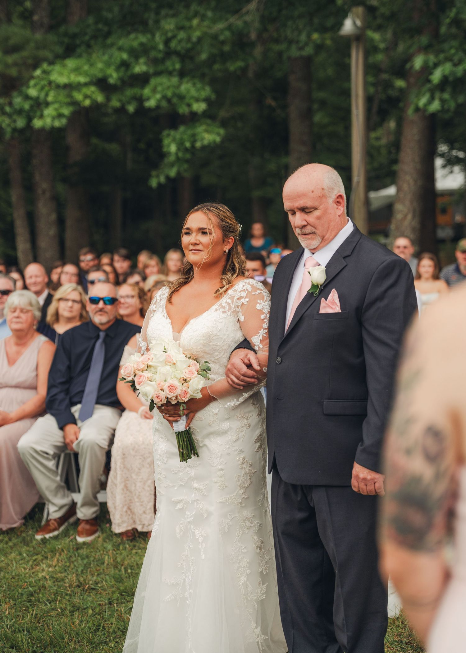 Bride in lace gown holds pink bouquet while being walked down the aisle by her father outdoors.