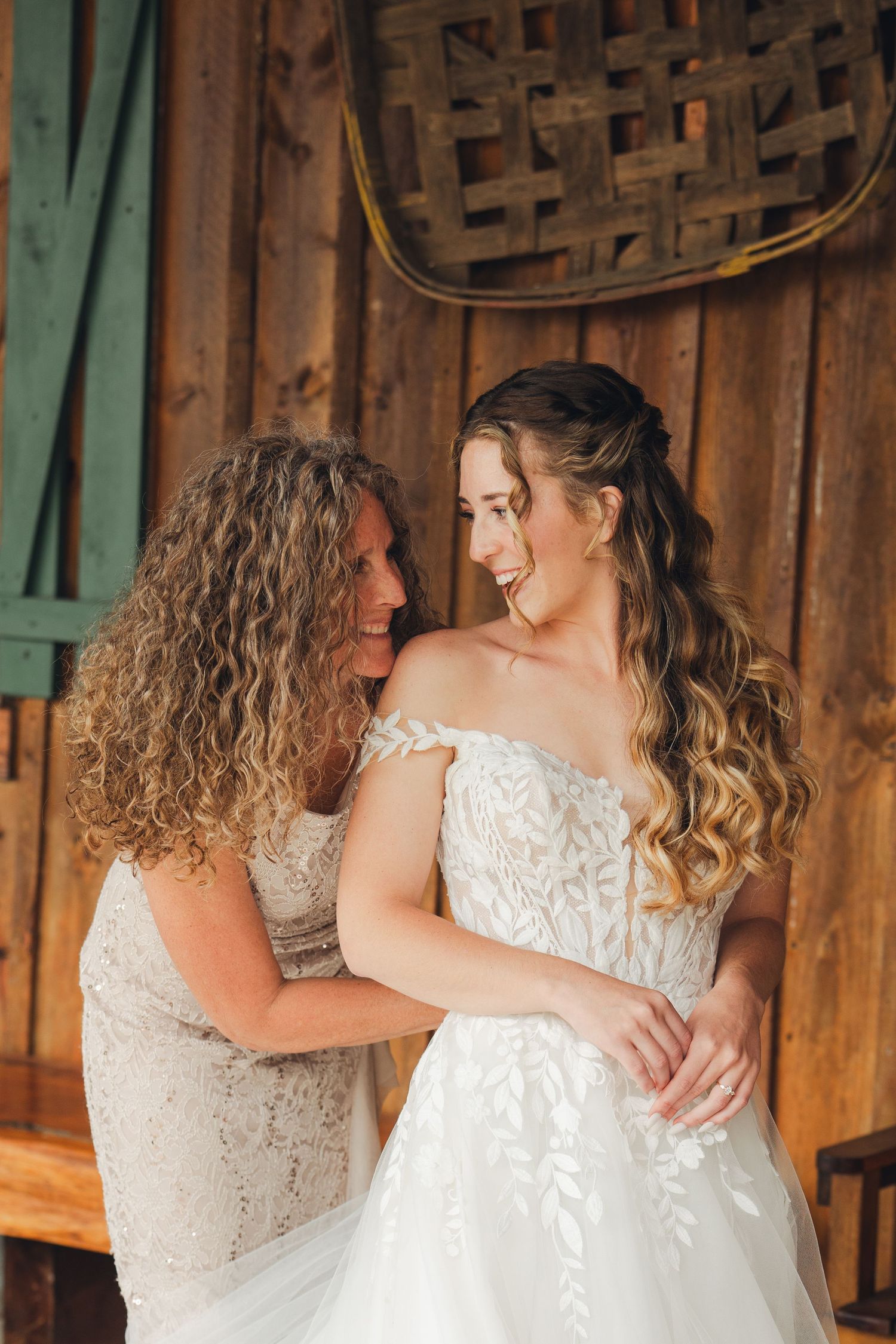 Mother helping bride into floral lace wedding gown in rustic barn setting.