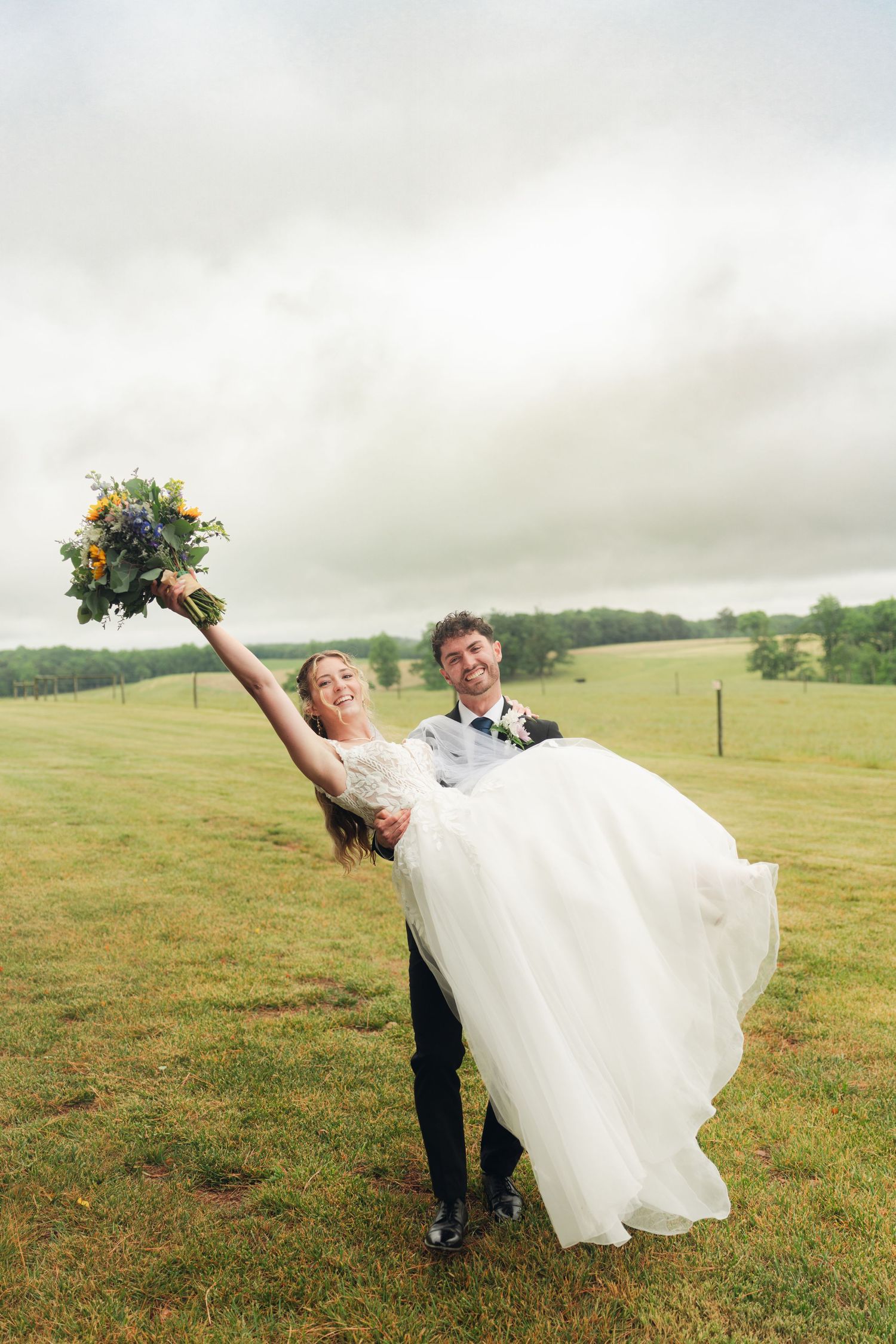 Groom lifting smiling bride in white gown as she raises bouquet in a lush green field.