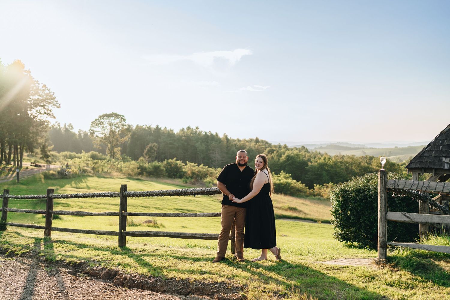 Smiling couple standing together by wooden fence with rolling green countryside and sunset behind them.