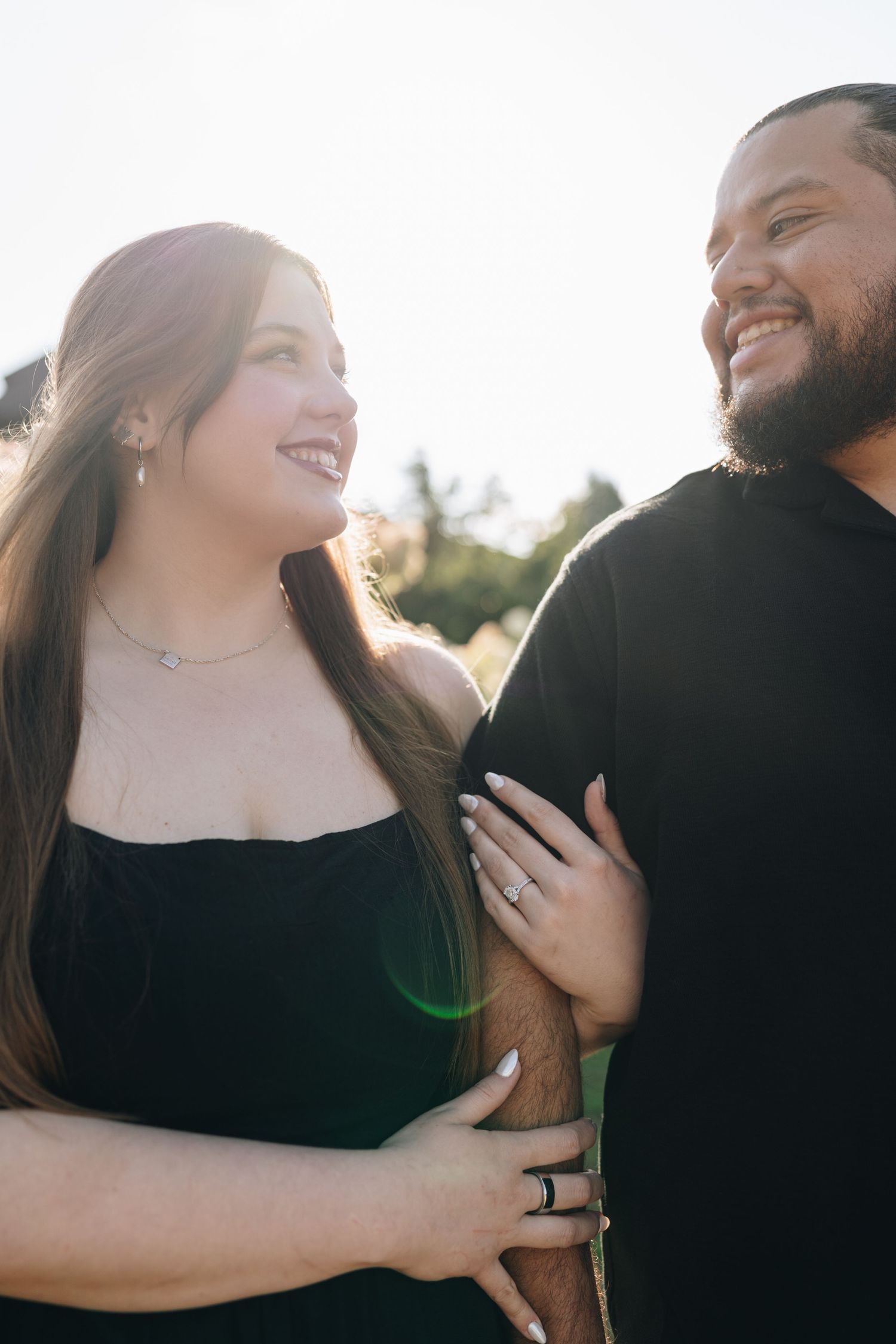 Smiling engaged couple embracing outdoors in bright sunlight, woman showing off her engagement ring.