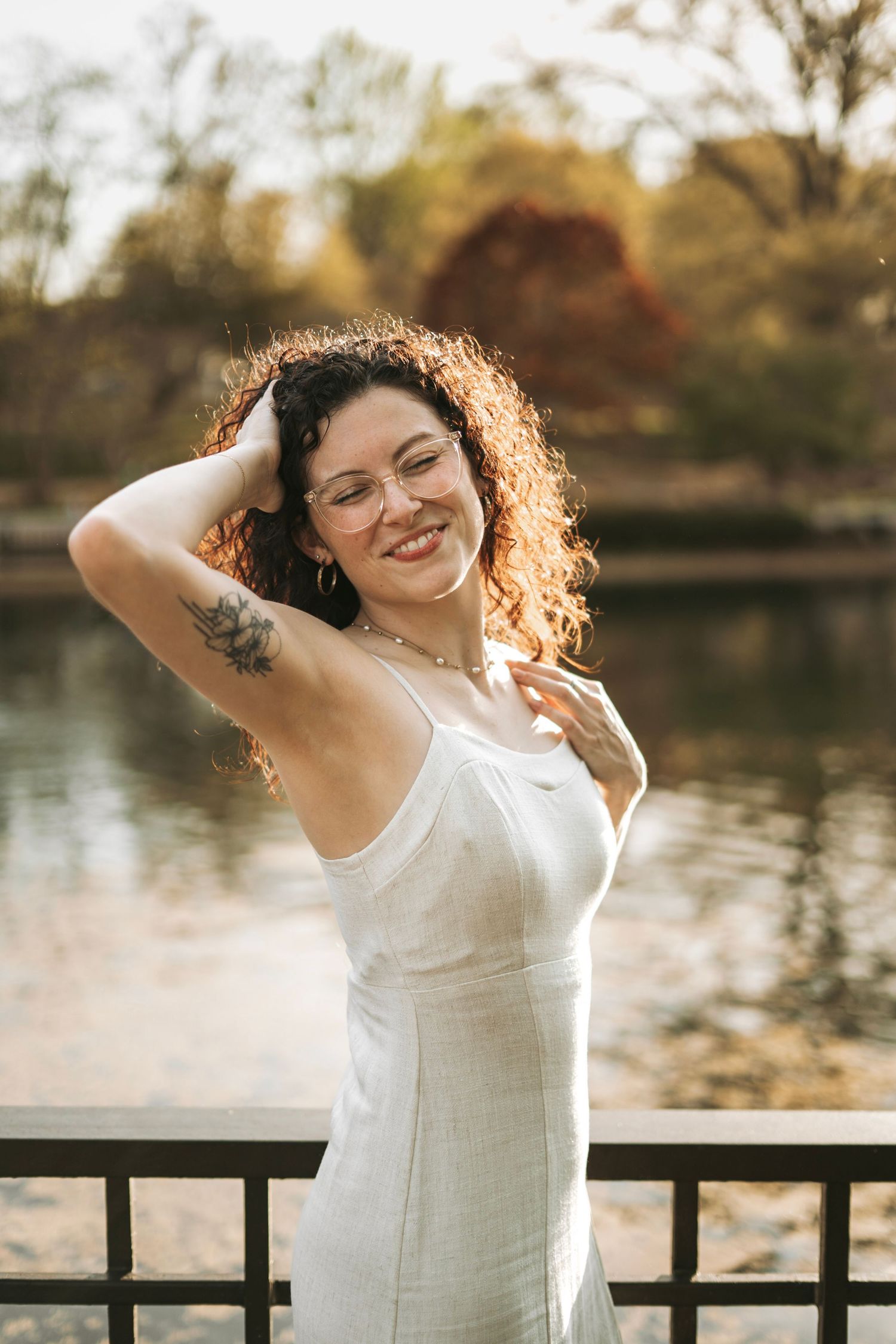 Smiling curly-haired woman in white dress with arm tattoo posing by sunlit lake railing.