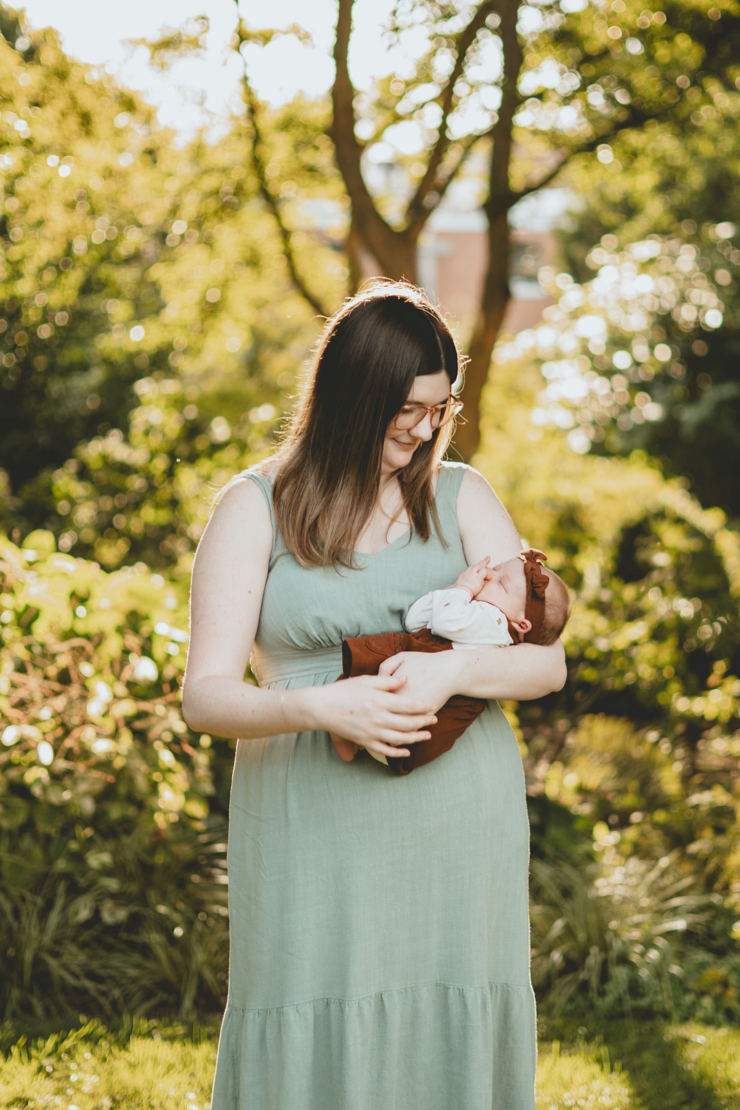 A woman in a sage green dress tenderly holds her sleeping newborn baby outdoors in a sunlit garden.