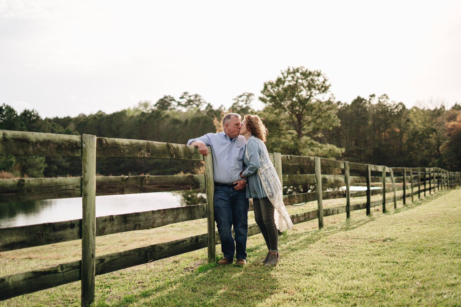 Couple leaning on a wooden fence by a peaceful pond surrounded by autumn trees at golden hour.