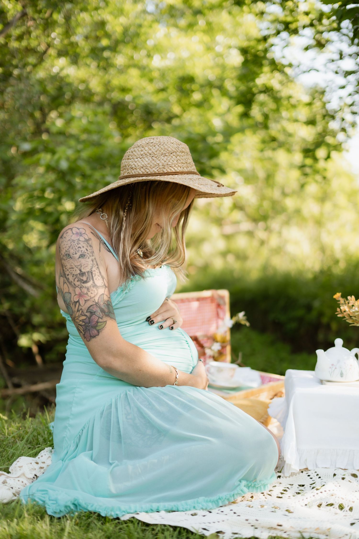 Tattooed pregnant woman in mint dress and straw hat sitting at outdoor picnic during maternity session.
