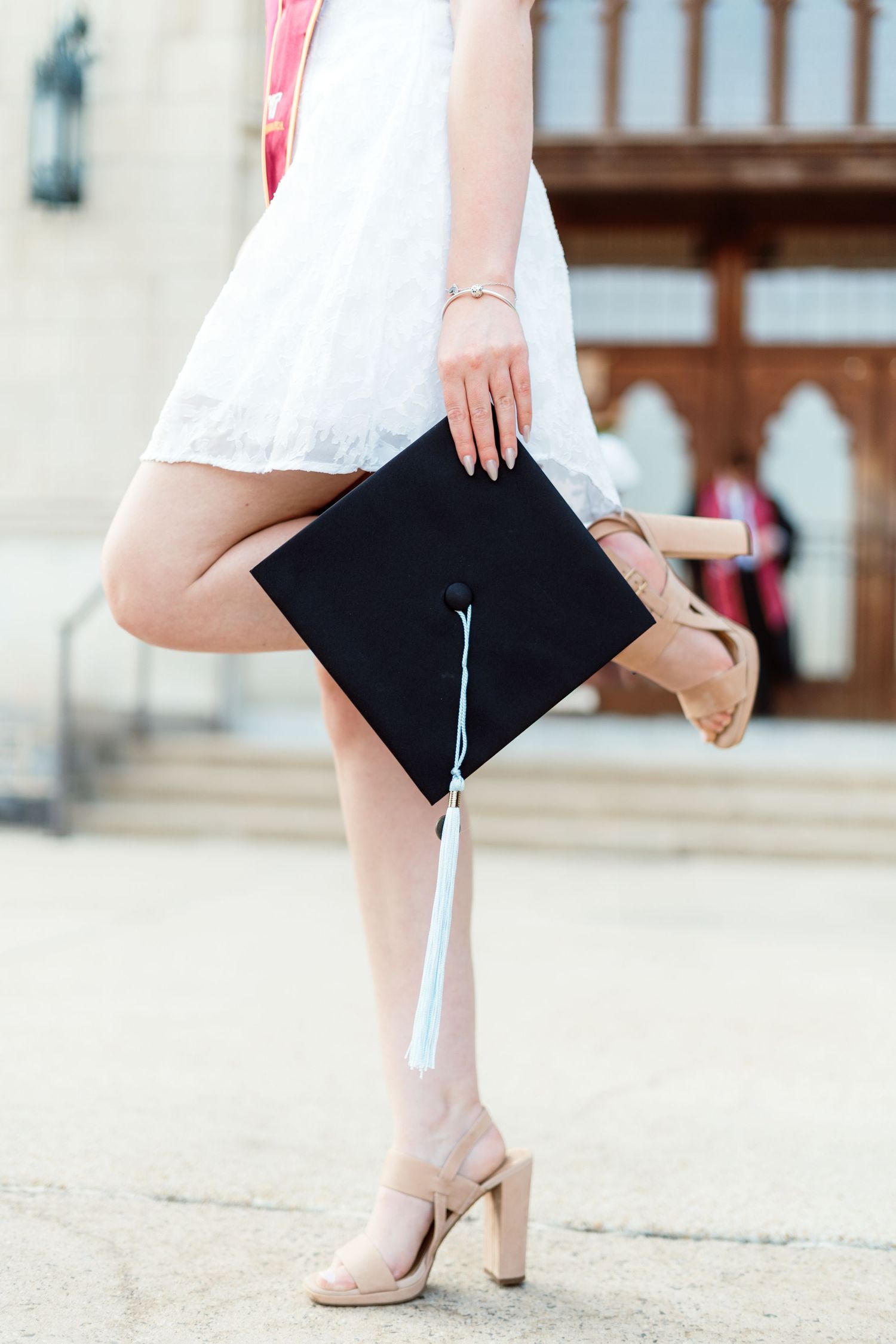 Close-up of graduate in white dress holding black mortarboard cap while posing in heels.