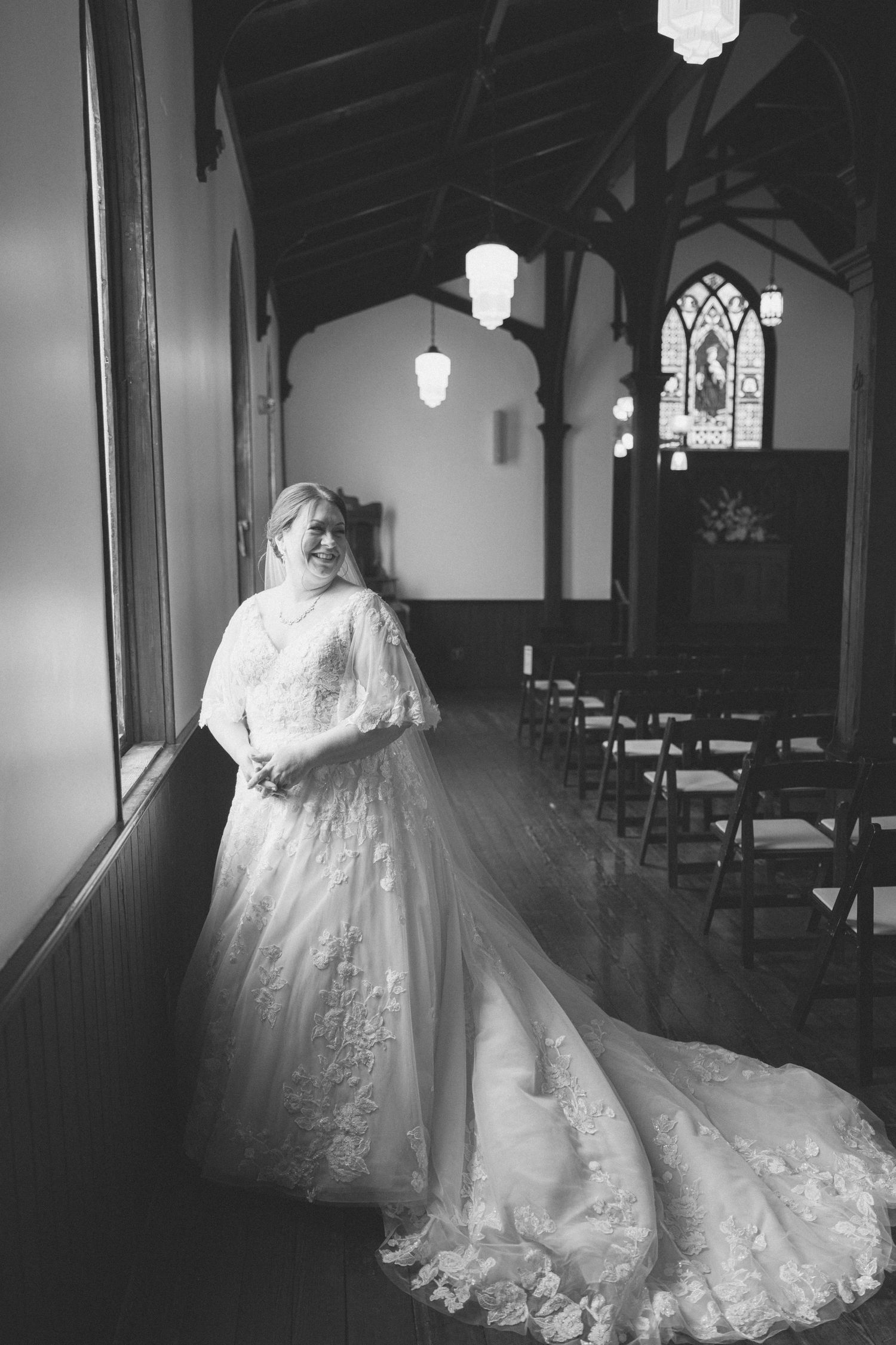 Black and white photo of bride in long lace gown with train posing inside historic church with stained glass.