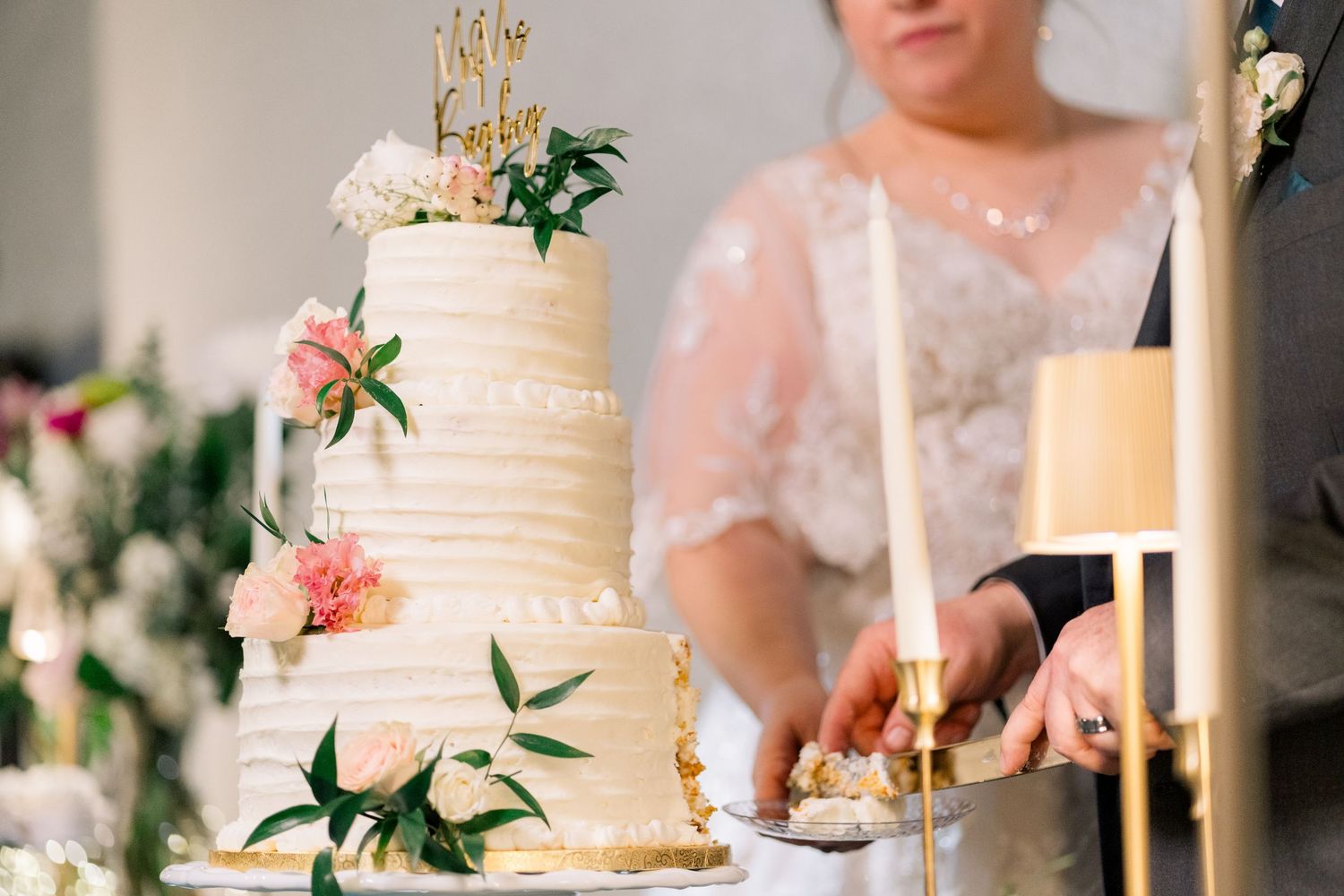 Three-tier white wedding cake with pink flowers and gold Mr and Mrs topper at reception.