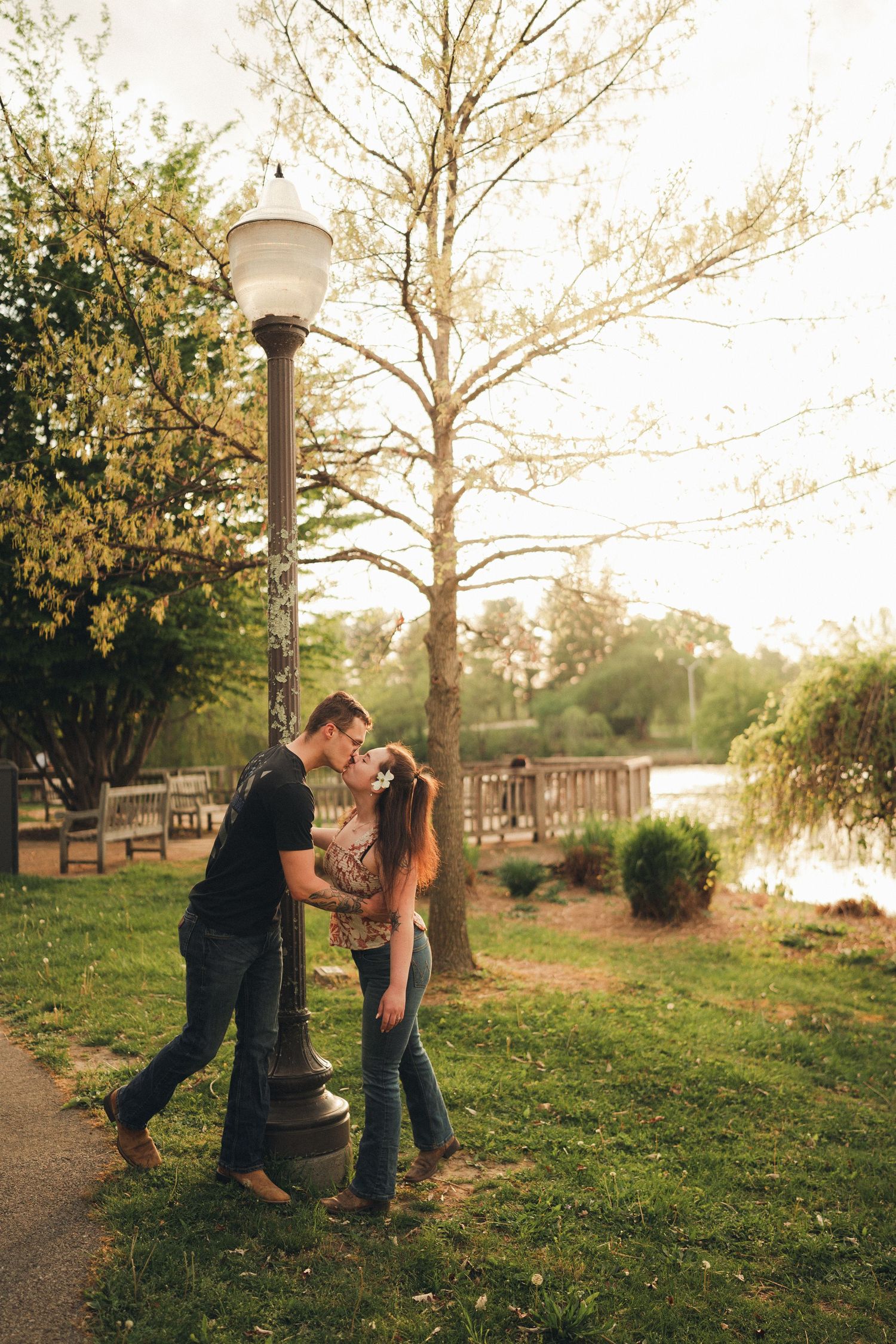 Romantic couple kissing by a lamppost in a park during sunset near a lake.