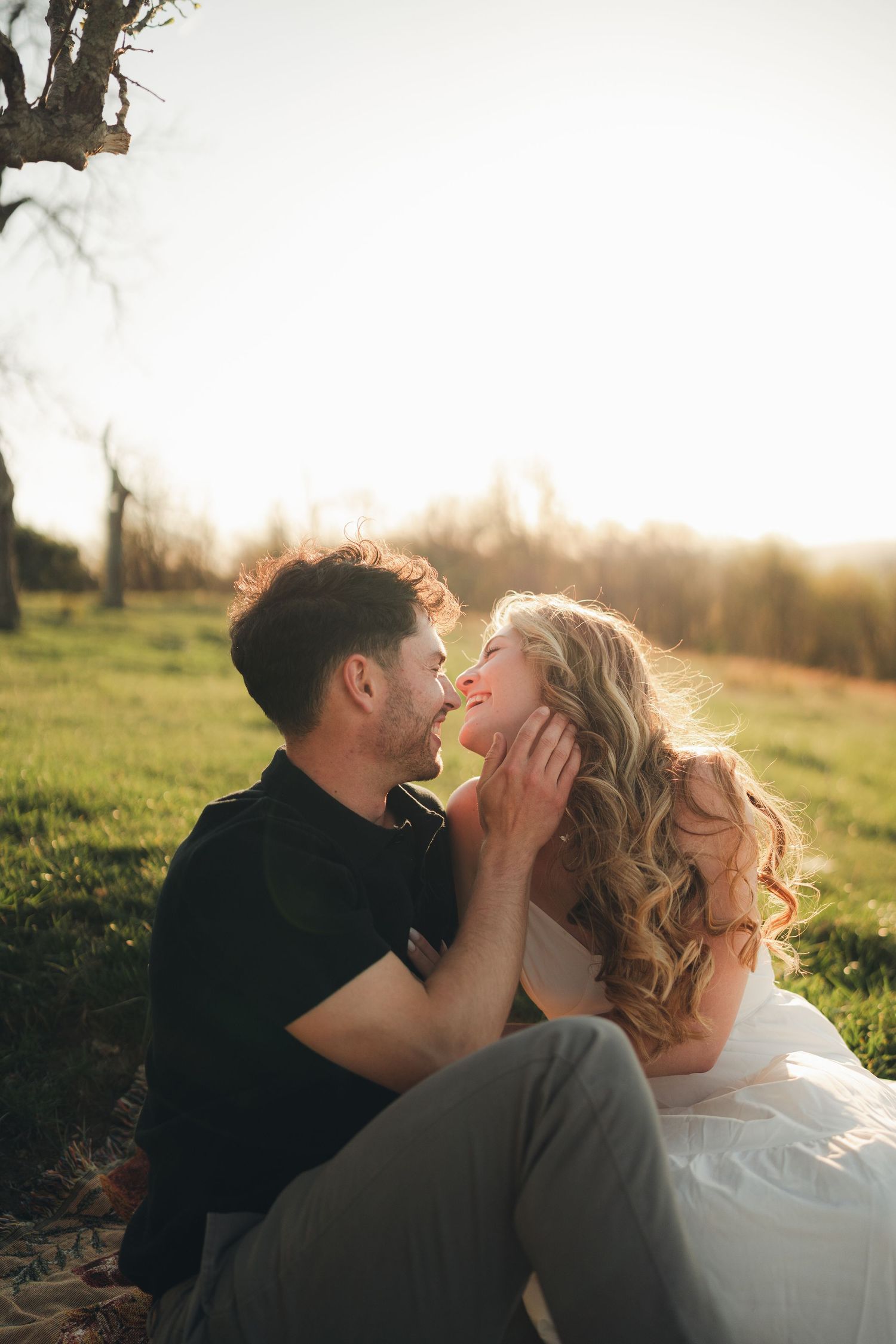 Couple sitting on grass laughing and almost kissing during golden sunset engagement photo session outdoors.