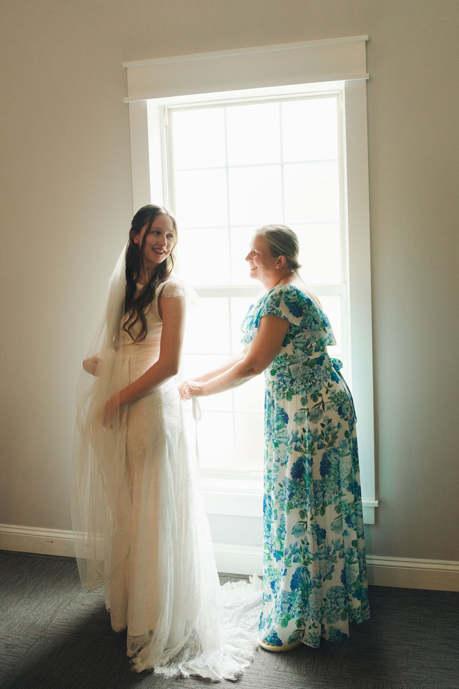 Bride in white gown smiling with woman in floral dress by sunlit window during wedding preparations.
