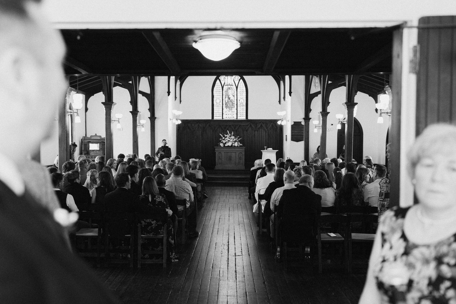 Black and white wide angle view of guests seated in a historic wooden church during a wedding ceremony.