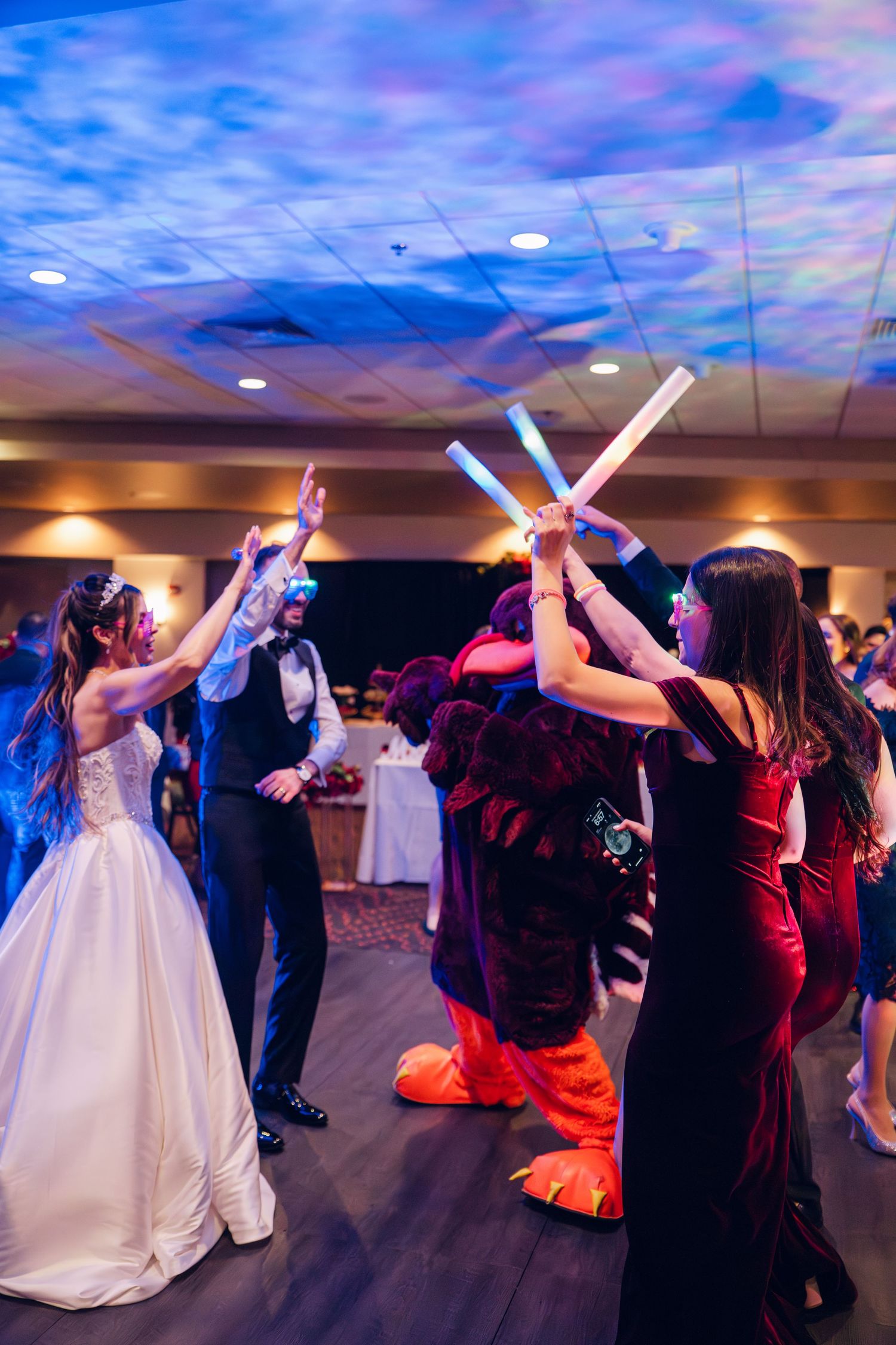Wedding guests dance with glowing foam sticks alongside mascot costume at lively reception.