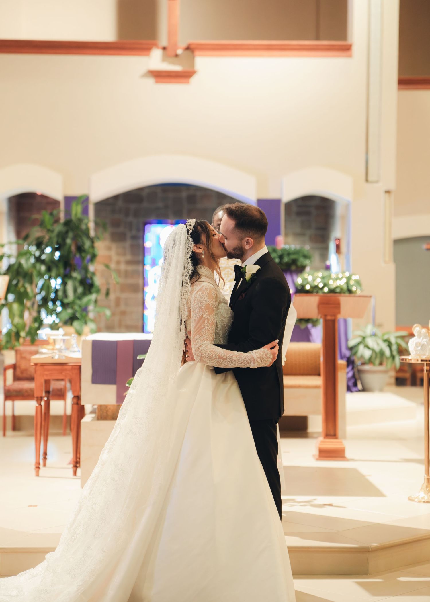 Newlyweds share romantic kiss at church altar in elegant wedding gown and black tuxedo.