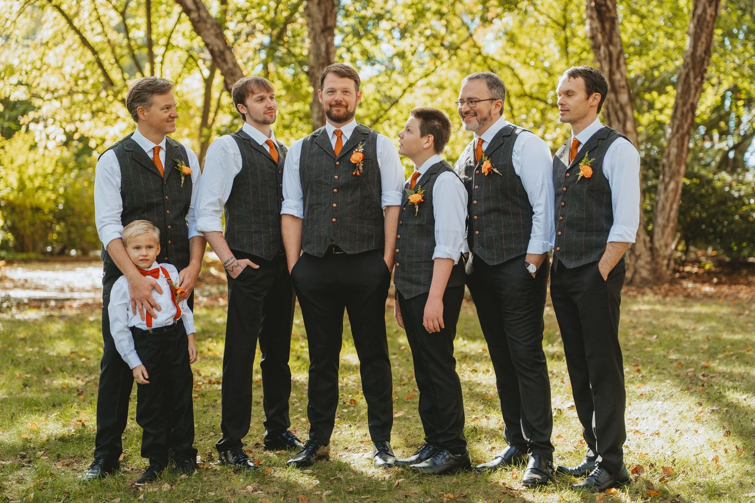 Groom and groomsmen in grey vests and orange ties posing outdoors at fall wedding.
