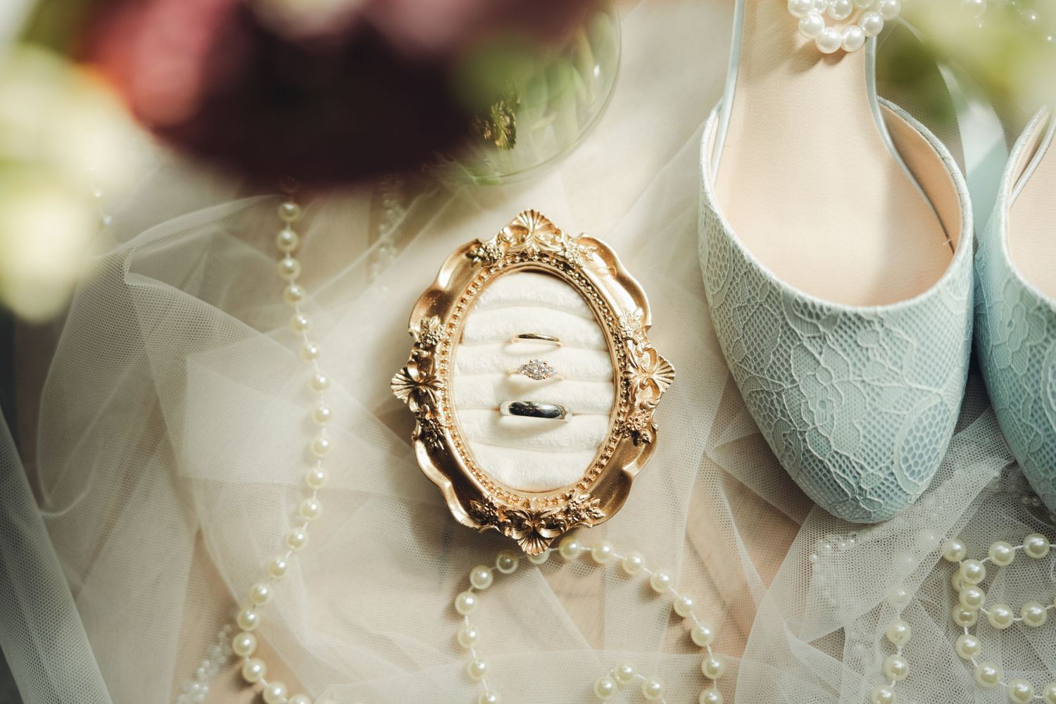 Gold ornate frame with wedding rings, pearl necklace, and mint lace bridal shoes on white tulle fabric.