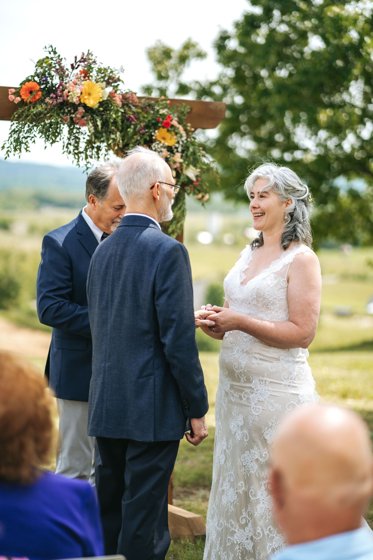 Older bride and groom exchanging vows at an outdoor ceremony beneath a colorful floral arch.