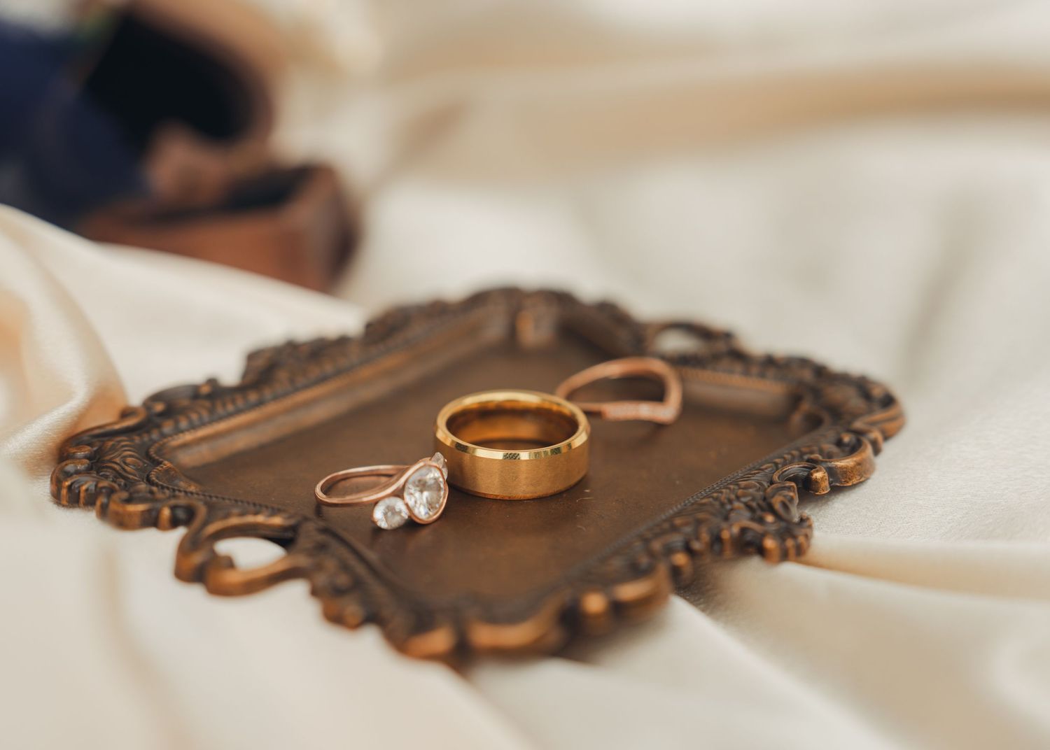 Gold wedding band and diamond engagement ring displayed on ornate vintage tray atop white satin fabric.