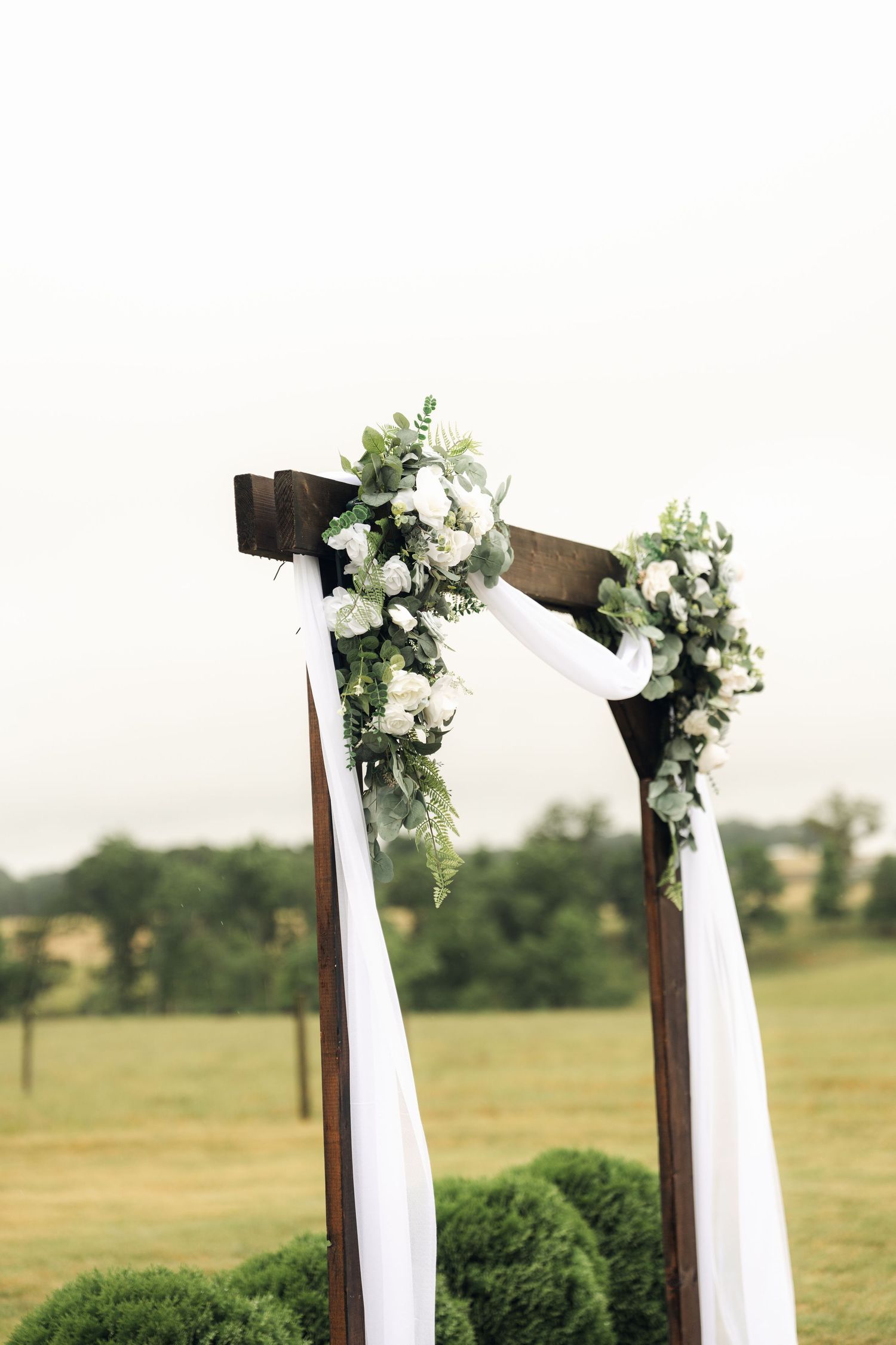 Rustic wooden wedding arch adorned with white roses, greenery, and flowing white fabric draping in an outdoor field setting.