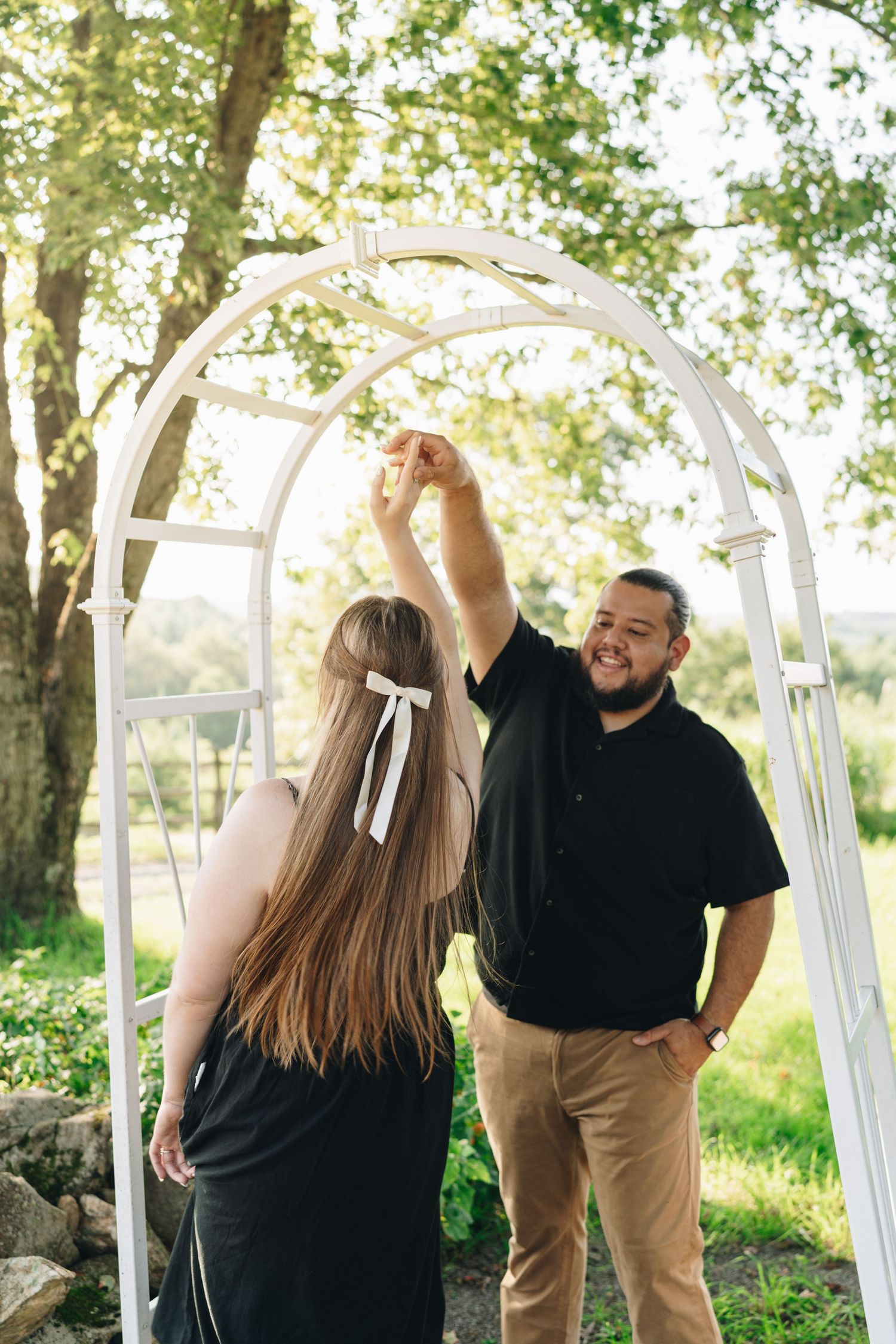 Joyful man spinning woman under a white garden arch surrounded by lush green trees.