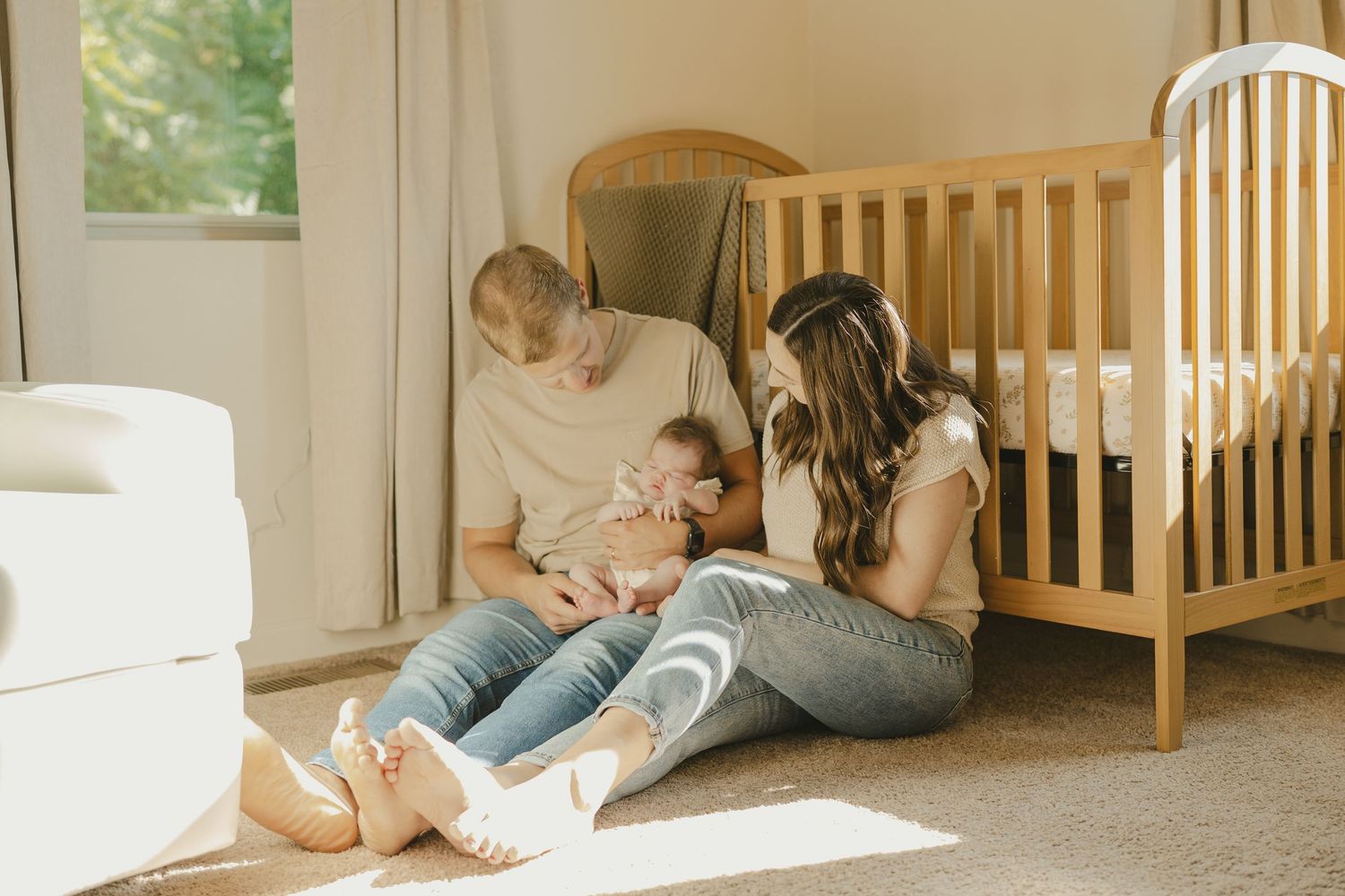 Parents sitting on nursery floor in sunlight caring for their newborn baby.