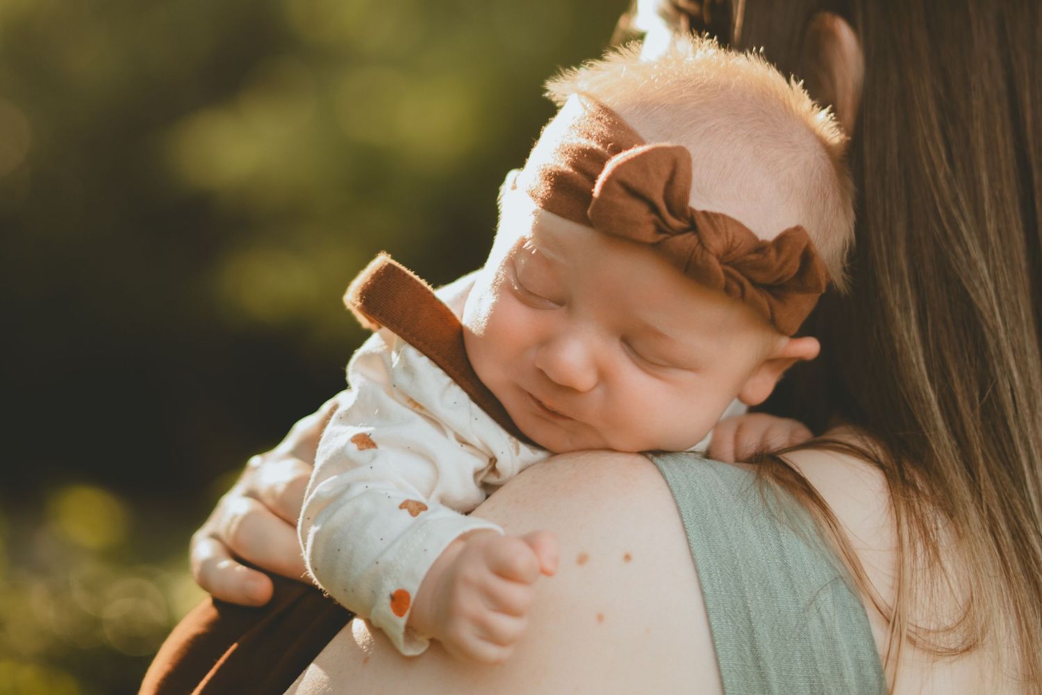 A sleeping newborn baby with a brown bow headband rests peacefully on a mothers shoulder outdoors.