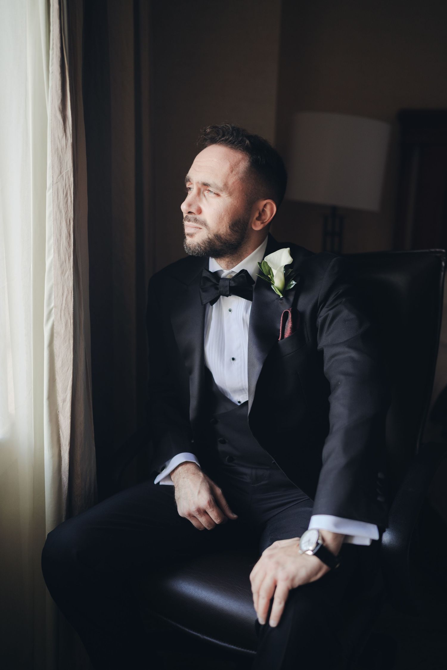 Elegant groom in black tuxedo with bow tie and floral boutonniere sitting by window on wedding day.