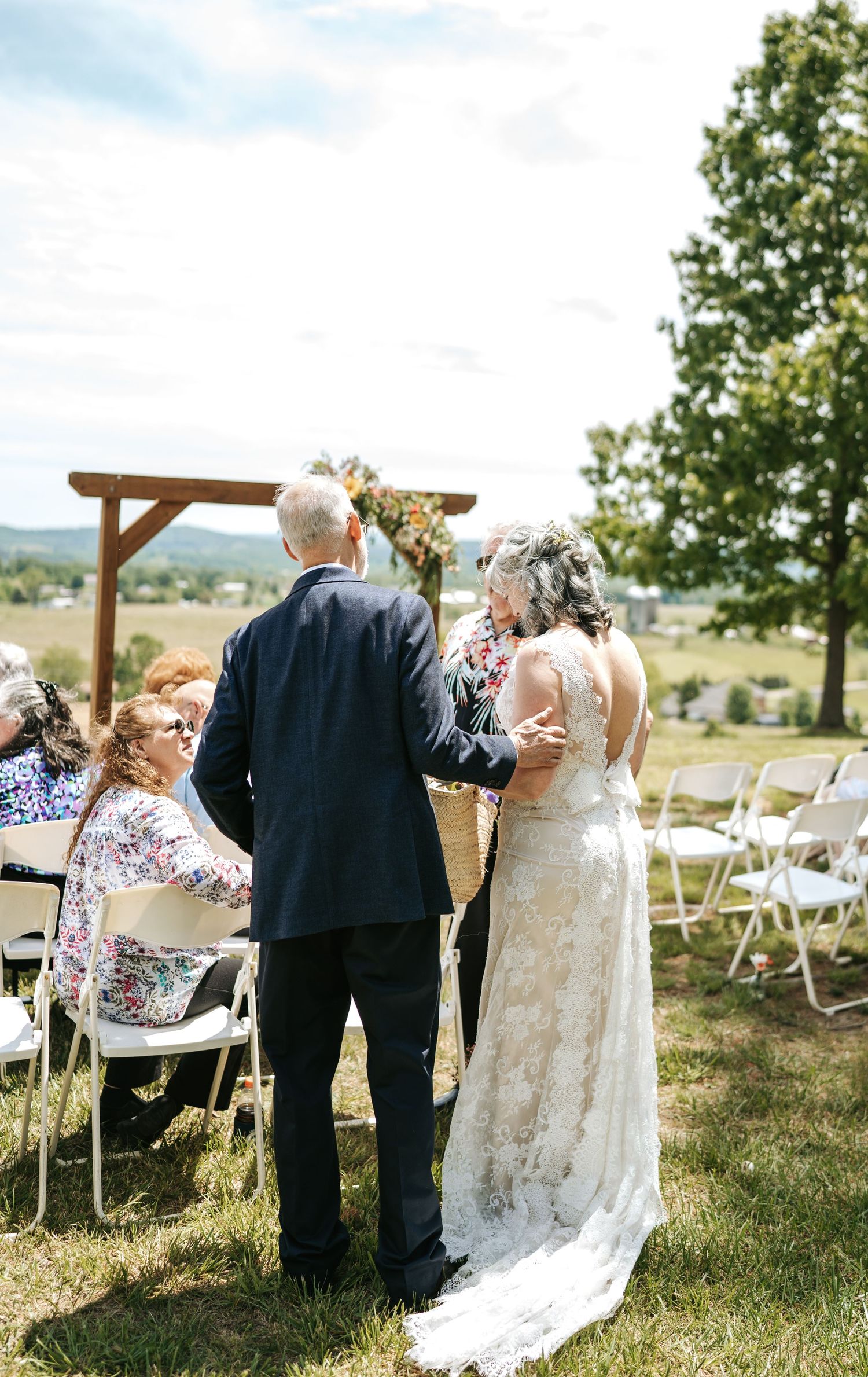 Bride and groom walk down outdoor aisle at scenic hillside wedding ceremony on sunny day.