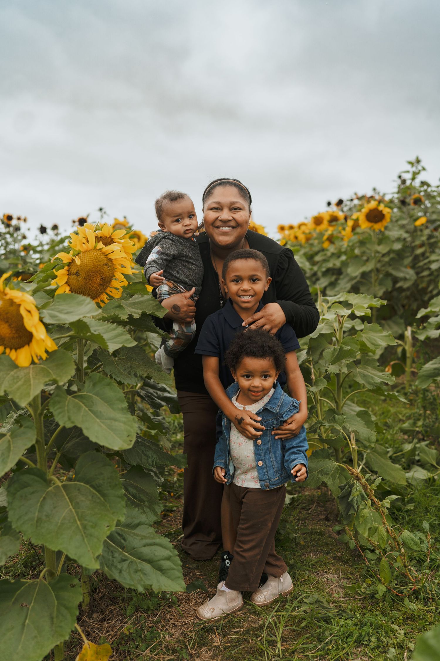 Smiling mother holding baby with two young children in a sunflower field.