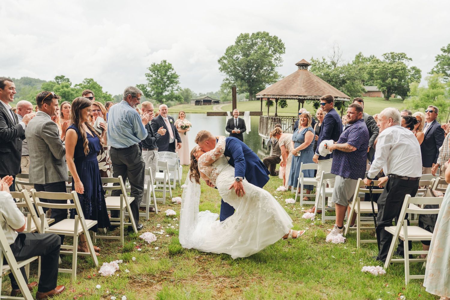 Groom dips bride for a kiss at outdoor wedding ceremony with guests cheering along flower petal-lined aisle near gazebo.