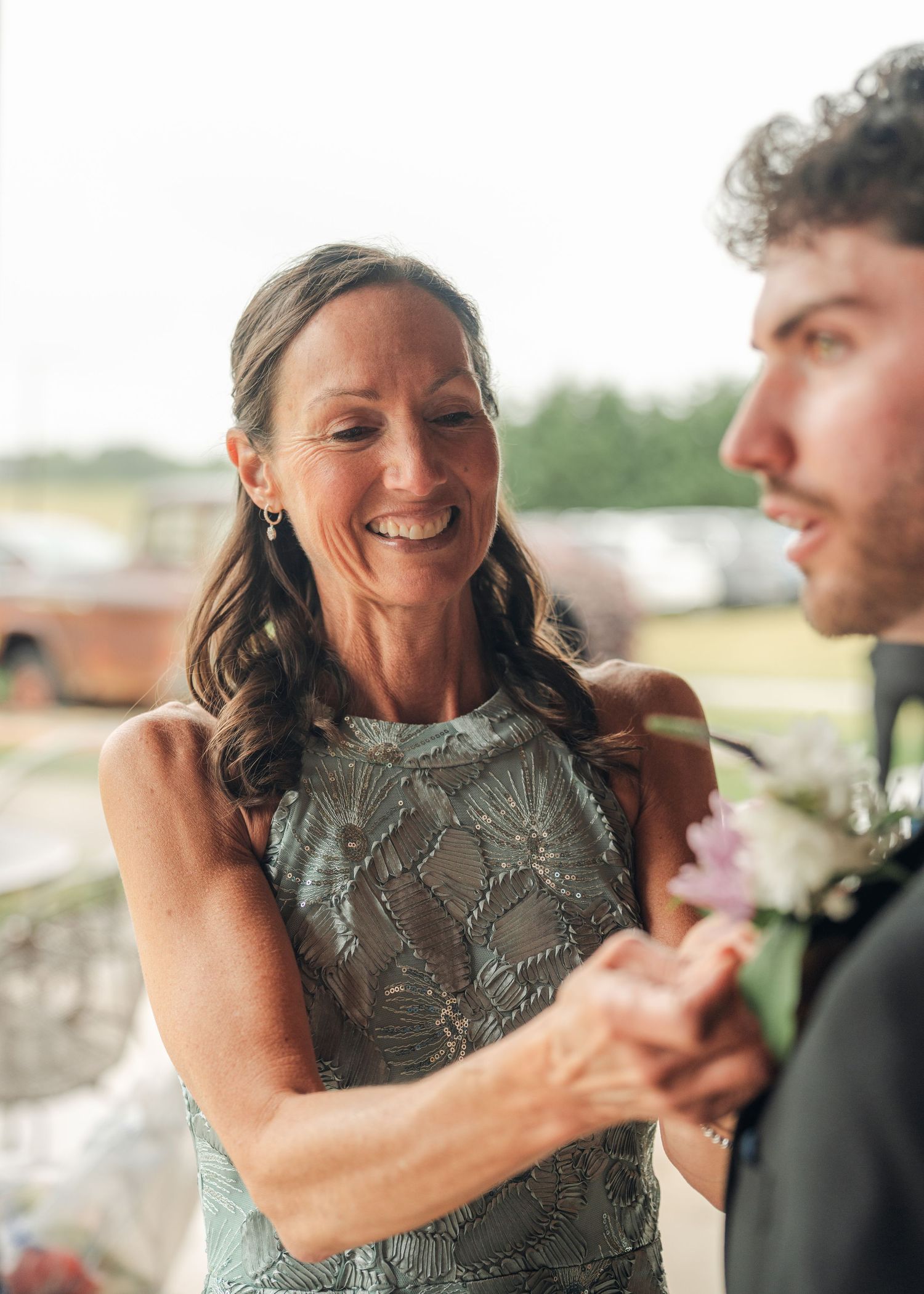 Mother of the groom smiling joyfully while pinning a boutonniere on her son at an outdoor wedding.