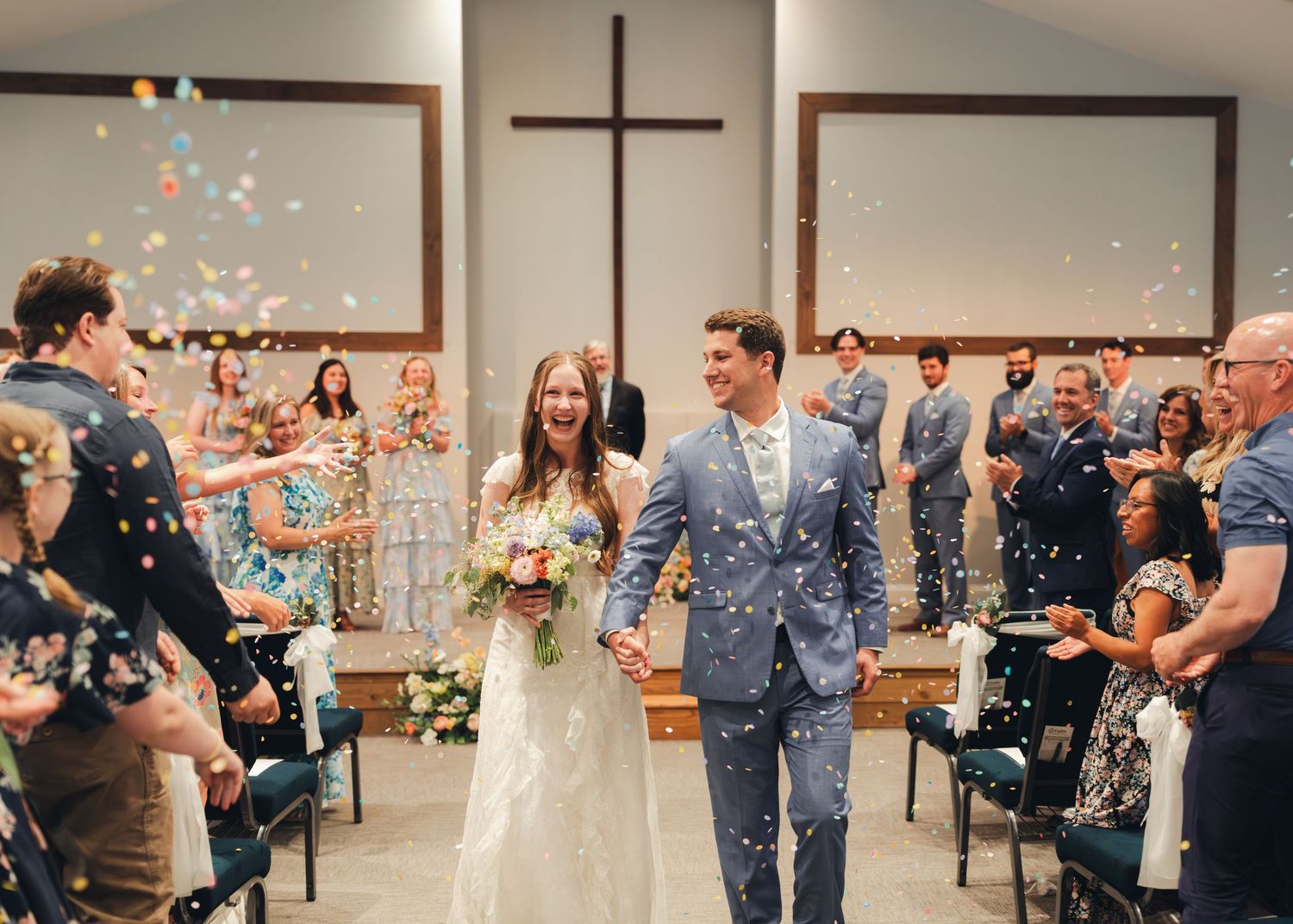 Joyful newlyweds walk down church aisle as guests toss colorful confetti during wedding recessional.