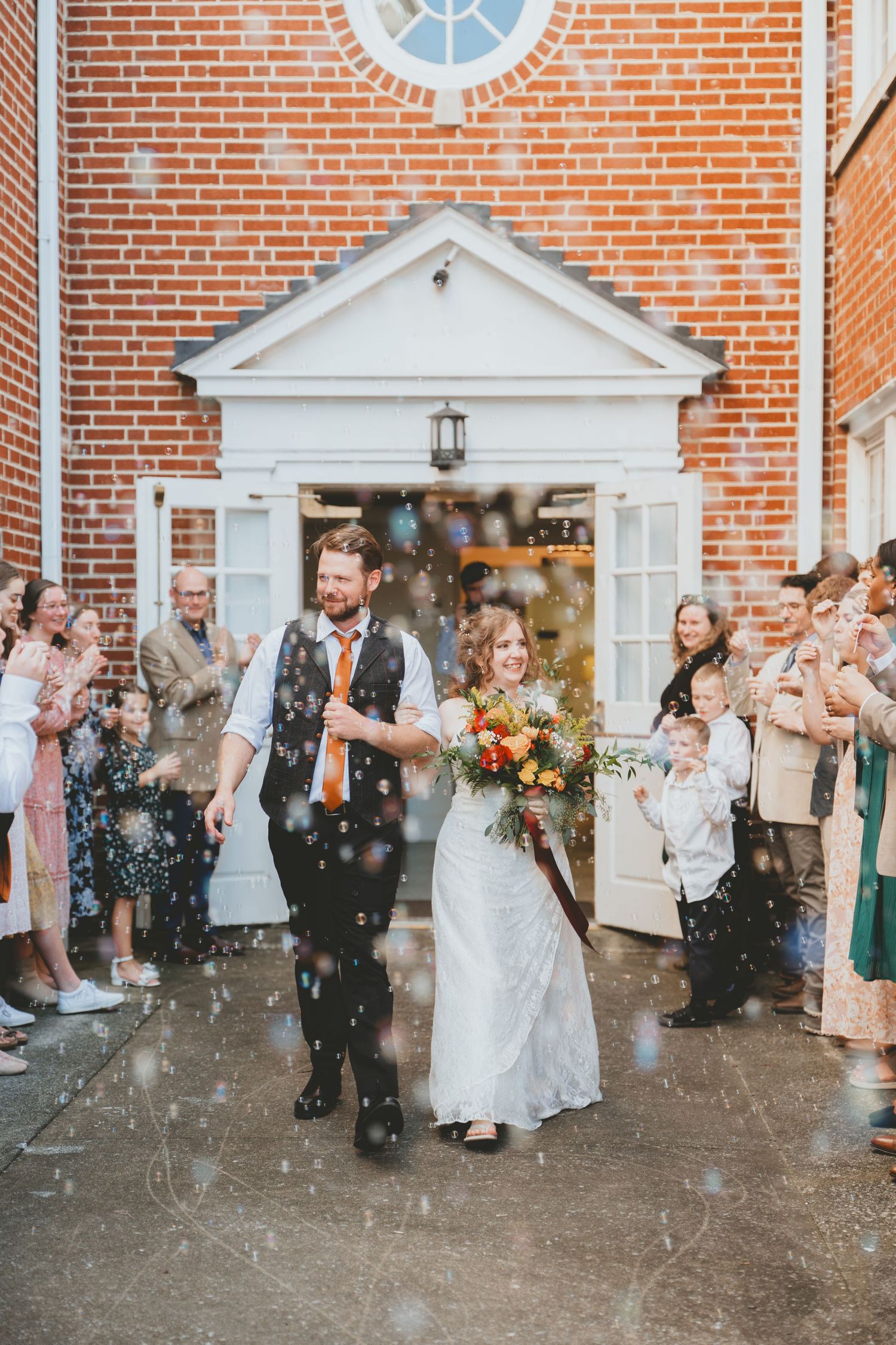 Newlyweds walk through a confetti send-off outside a classic brick church, bride carrying colorful fall flowers.