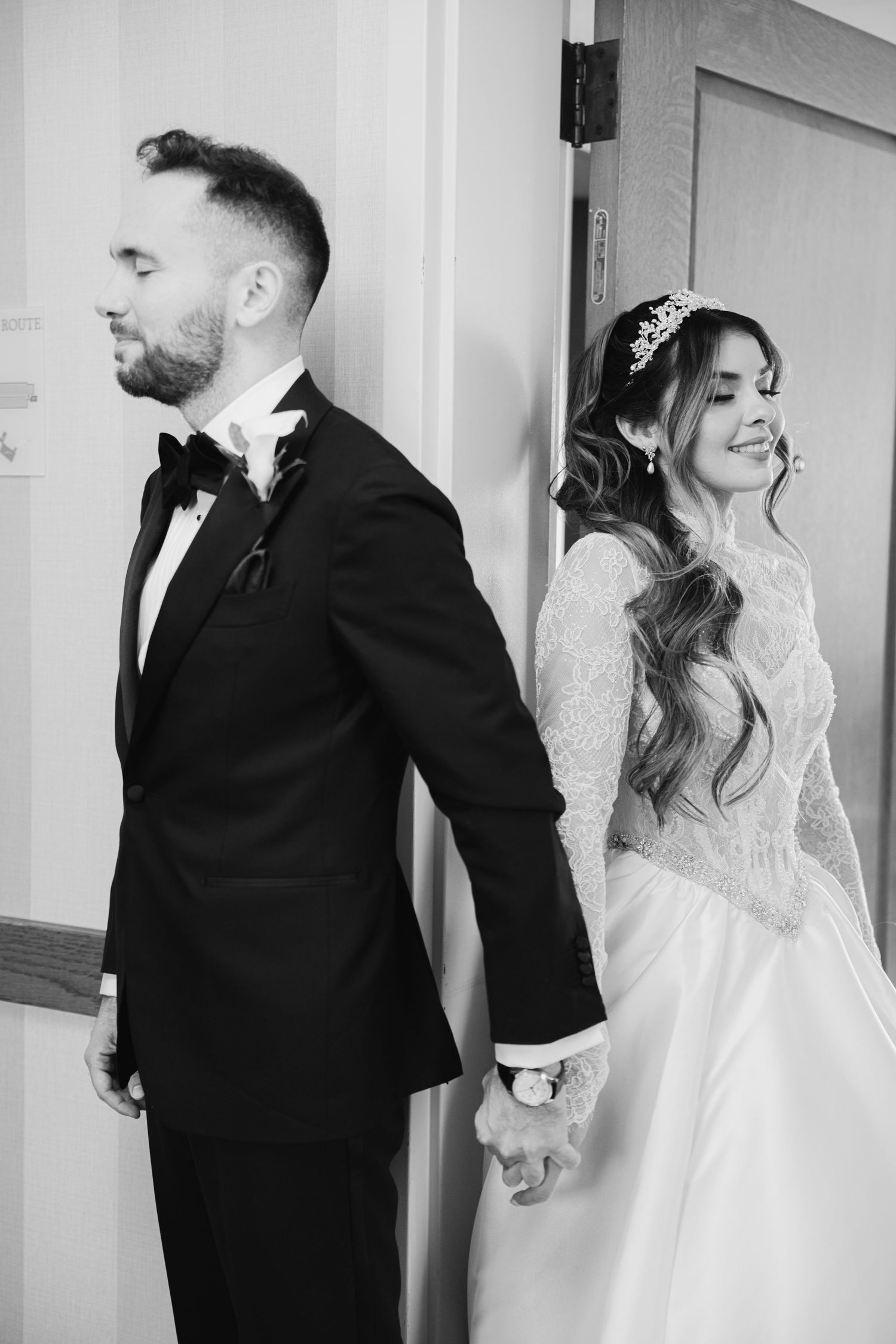 Black and white wedding portrait of bride and groom holding hands back to back by a door.