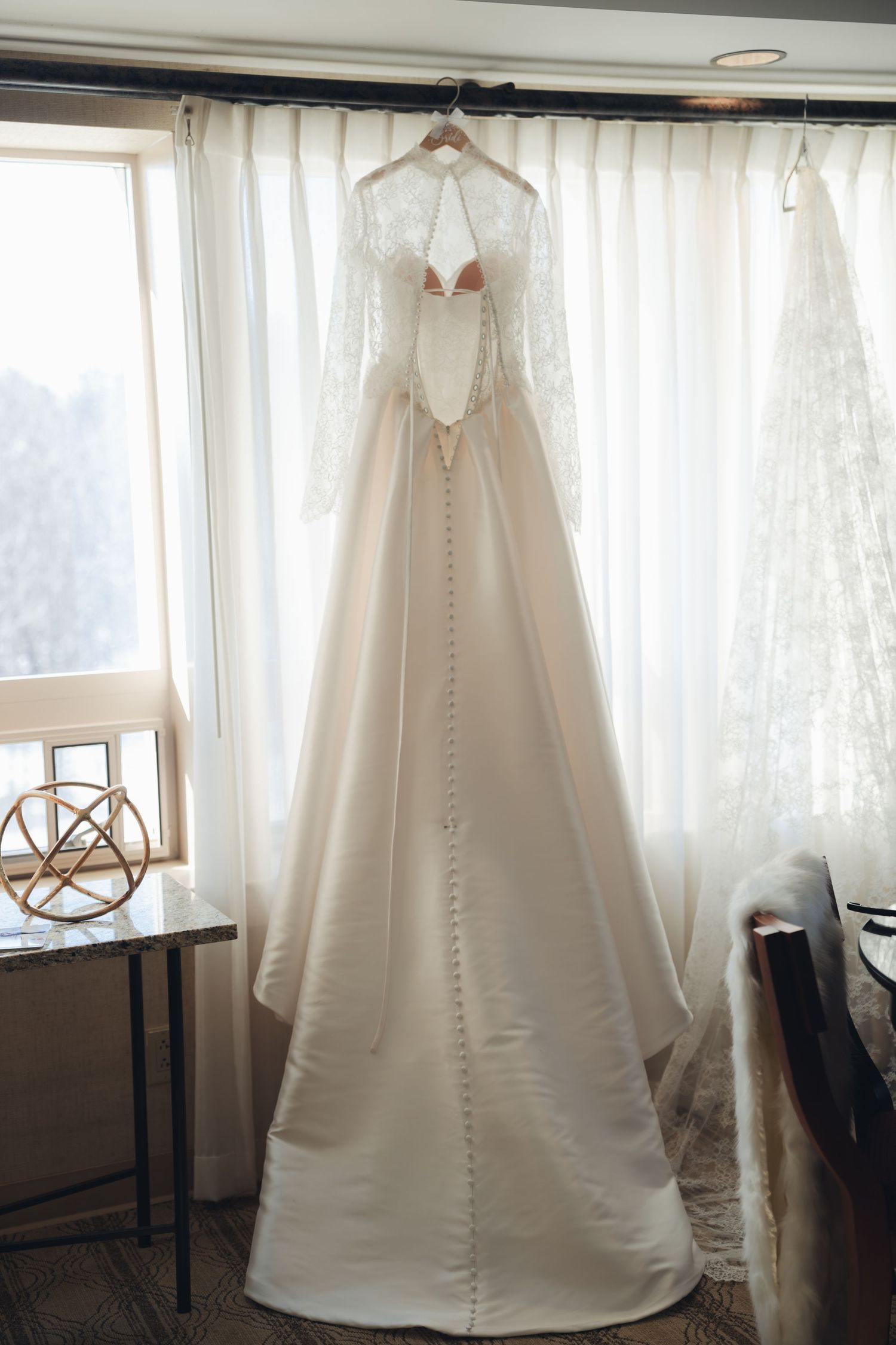 Elegant long-sleeve lace and satin A-line wedding dress hanging by window on hanger.