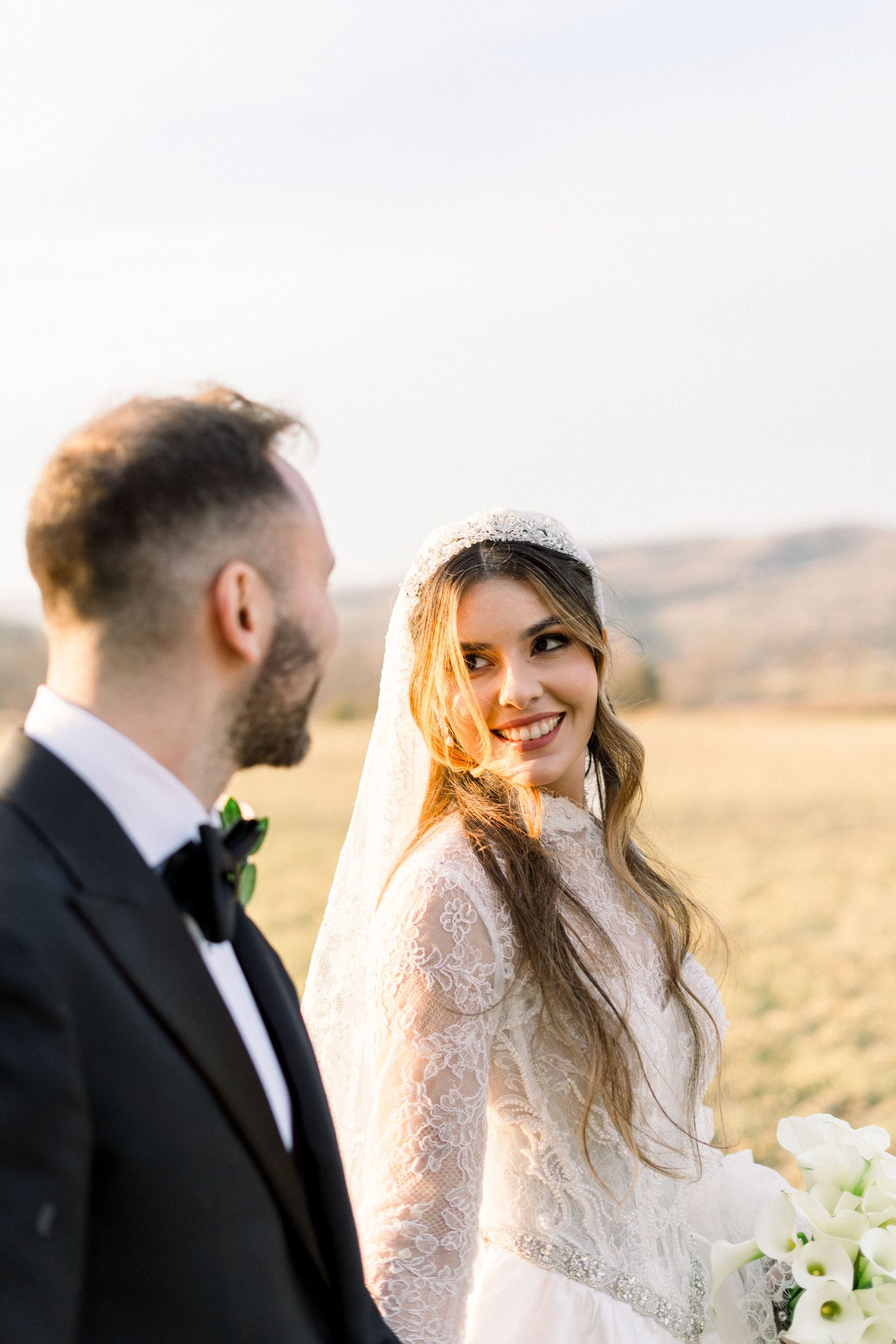 Radiant bride in lace gown and tiara smiles at groom in black tuxedo in sunny outdoor field.