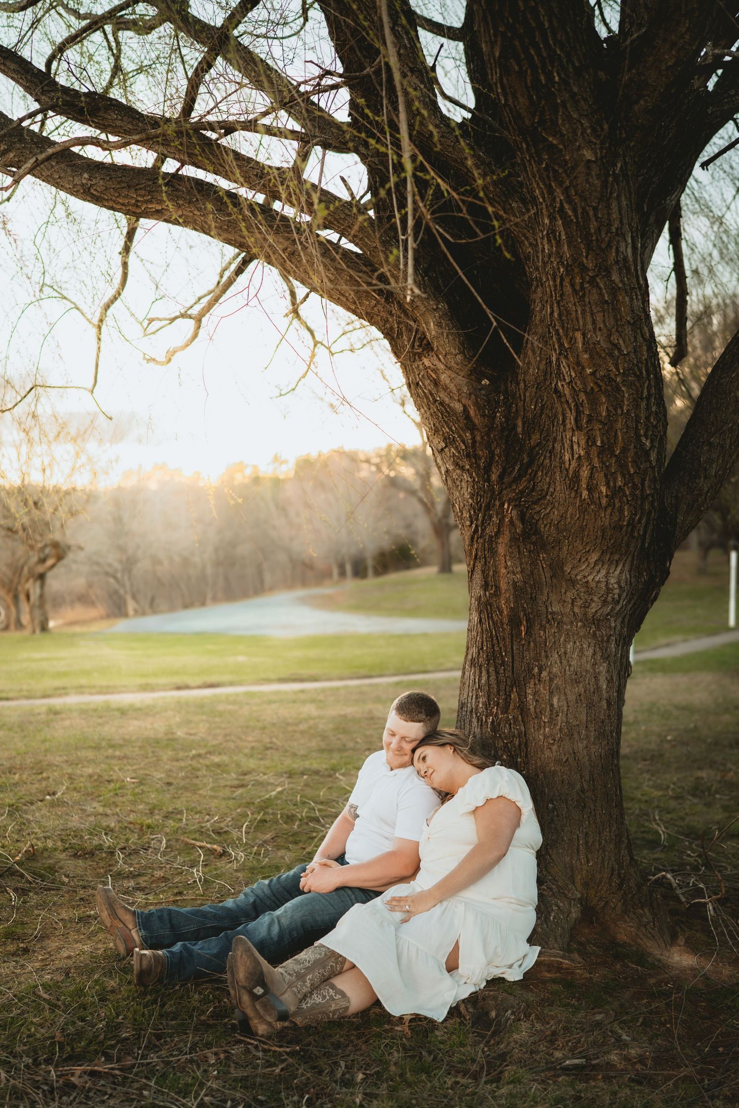 Couple sitting under large tree at golden hour, man embracing pregnant woman in white dress outdoors.