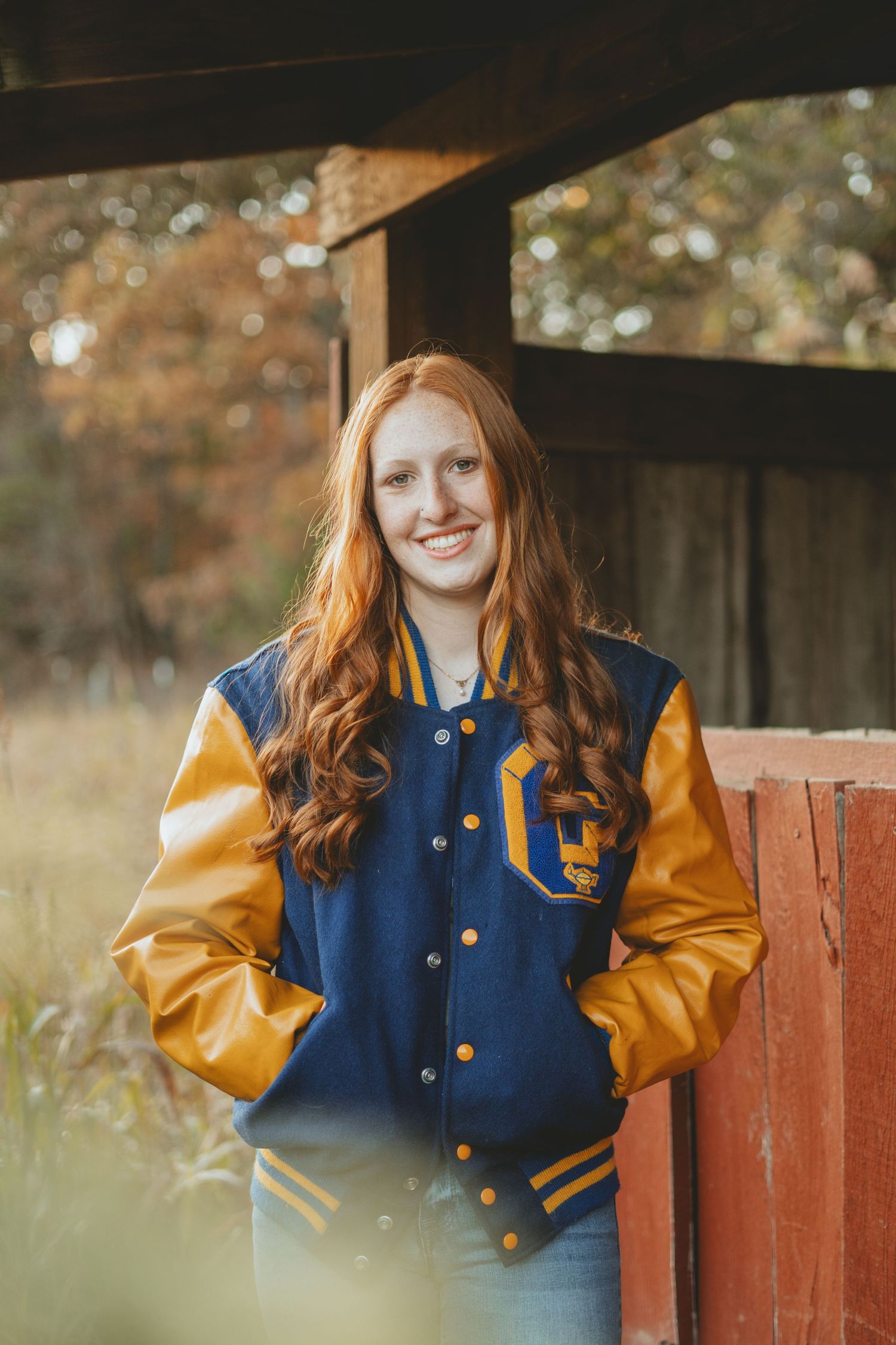 Smiling teenage girl with red hair wearing a navy and gold varsity jacket leaning against a red barn post in autumn.
